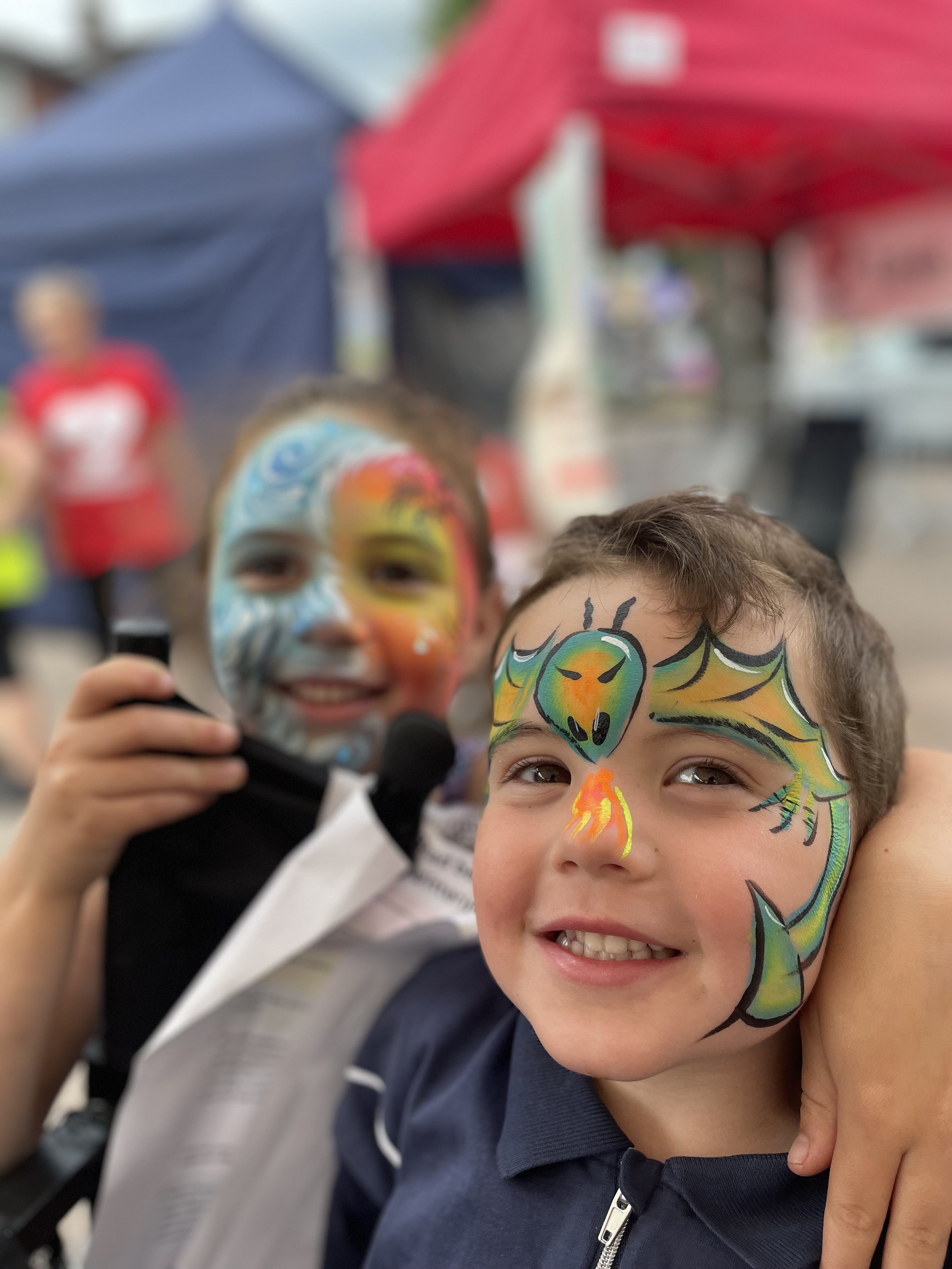 Two smiling children showcase vibrant face painting, with colorful designs against a backdrop of festive tents, embodying joyful event vibes.