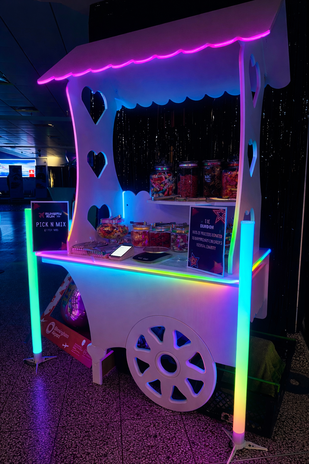 A vibrant candy cart displays colorful jars filled with sweets, illuminated by neon lights, set against a shimmering, dark backdrop.