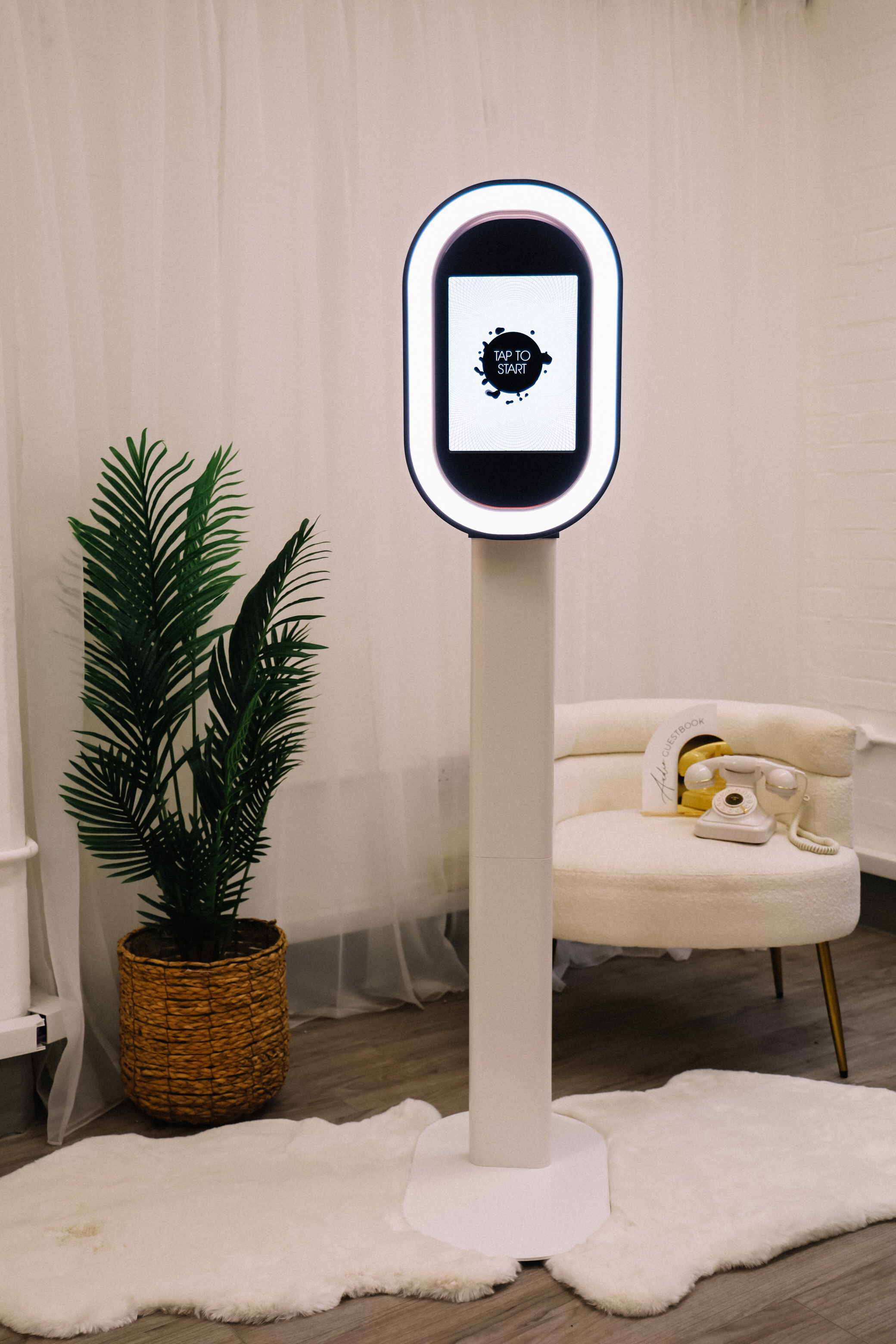 A sleek white photo booth stands against a soft, draped backdrop, complemented by a lush green plant and cozy golden-accented furniture, creating a modern, inviting atmosphere.