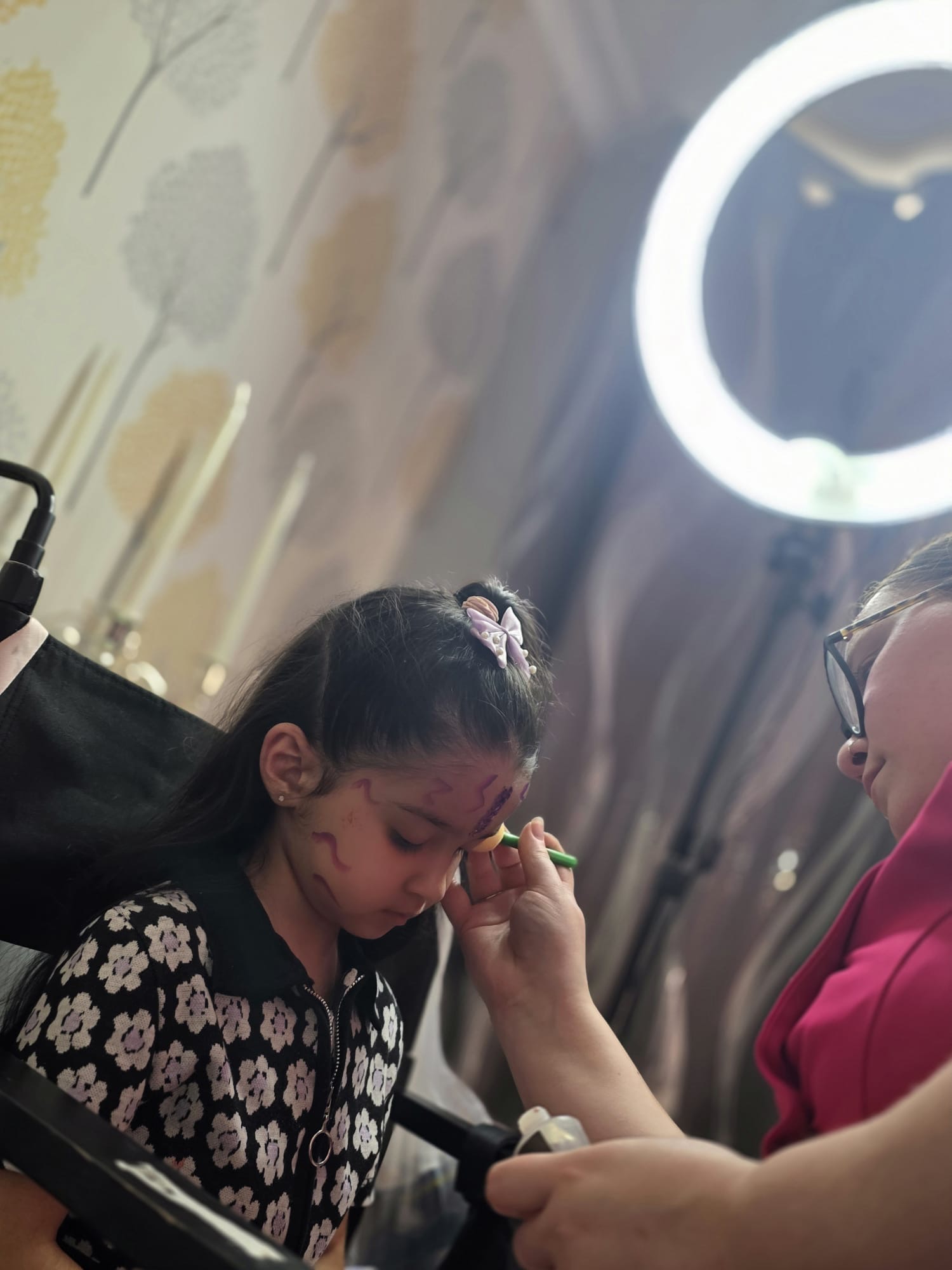 A young girl with floral-patterned attire sits in a black chair, focused on face painting. Soft lighting enhances the vibrant colors.