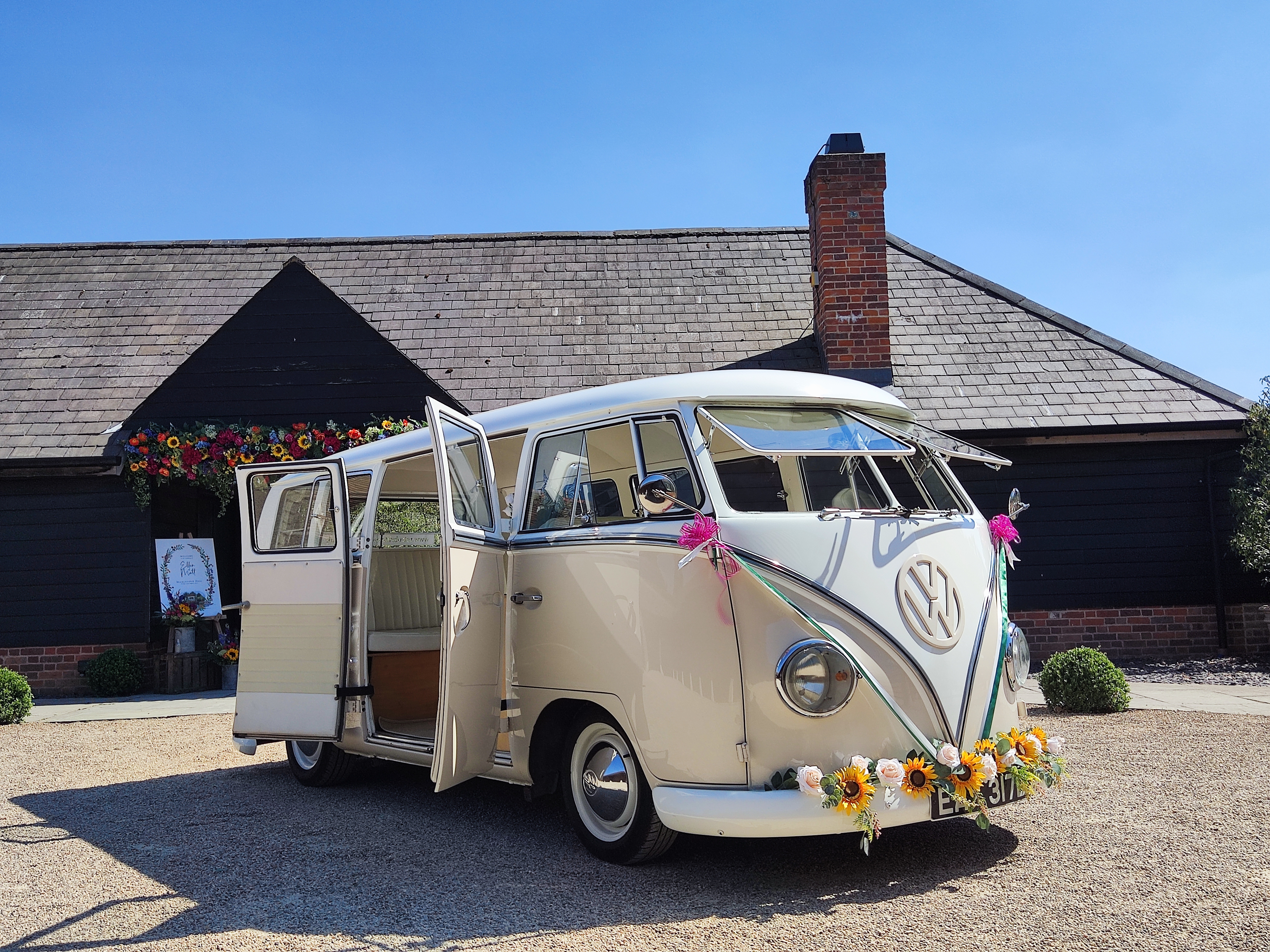 A vintage VW van adorned with vibrant flowers stands in front of a rustic black barn, basking in bright daylight, creating a charming atmosphere.