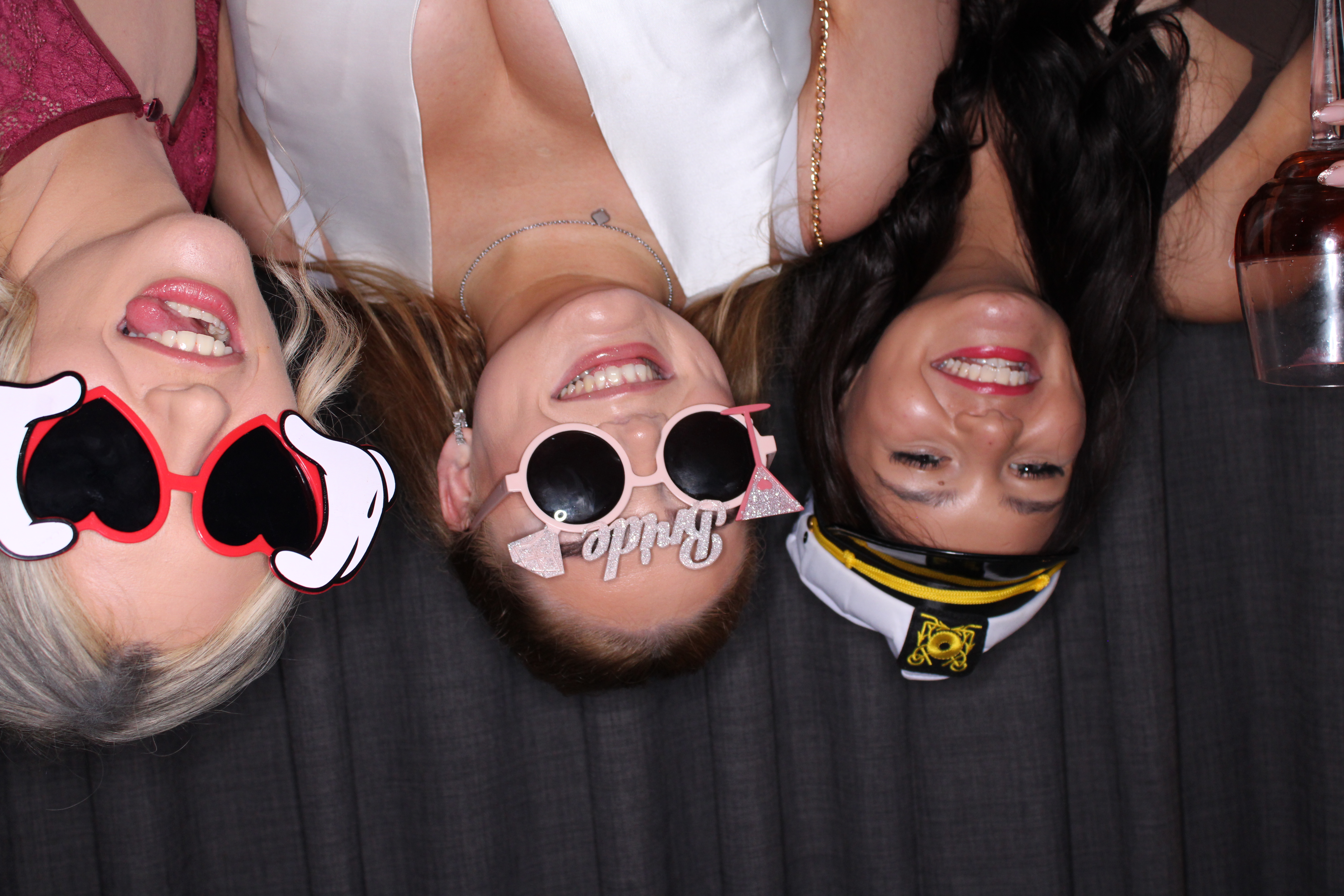 Three women smile in a photo booth, wearing bridal party props and heart sunglasses, with warm flash lighting and party vibes.