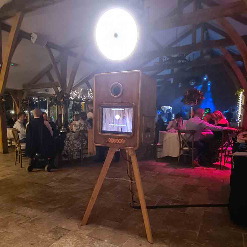 A vintage photo booth with warm wood tones stands on sturdy legs, illuminated by soft halo lighting, while guests enjoy a lively evening.
