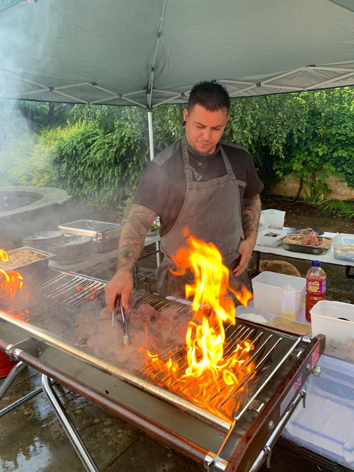 A chef skillfully grills marinated meats over an open flame, with smoke swirling around a green tent, creating a vibrant, lively atmosphere.