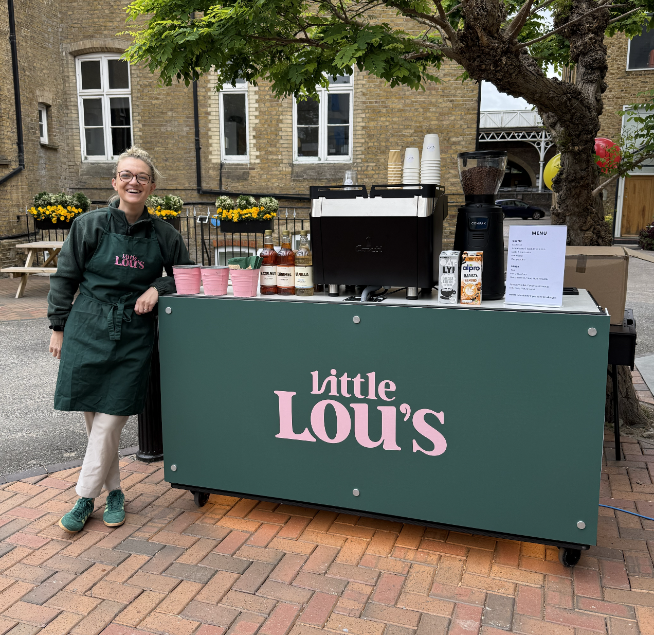 Smiling barista beside Little Lou’s coffee catering cart, with polished espresso machine, syrups, cups and menu in soft daylight.