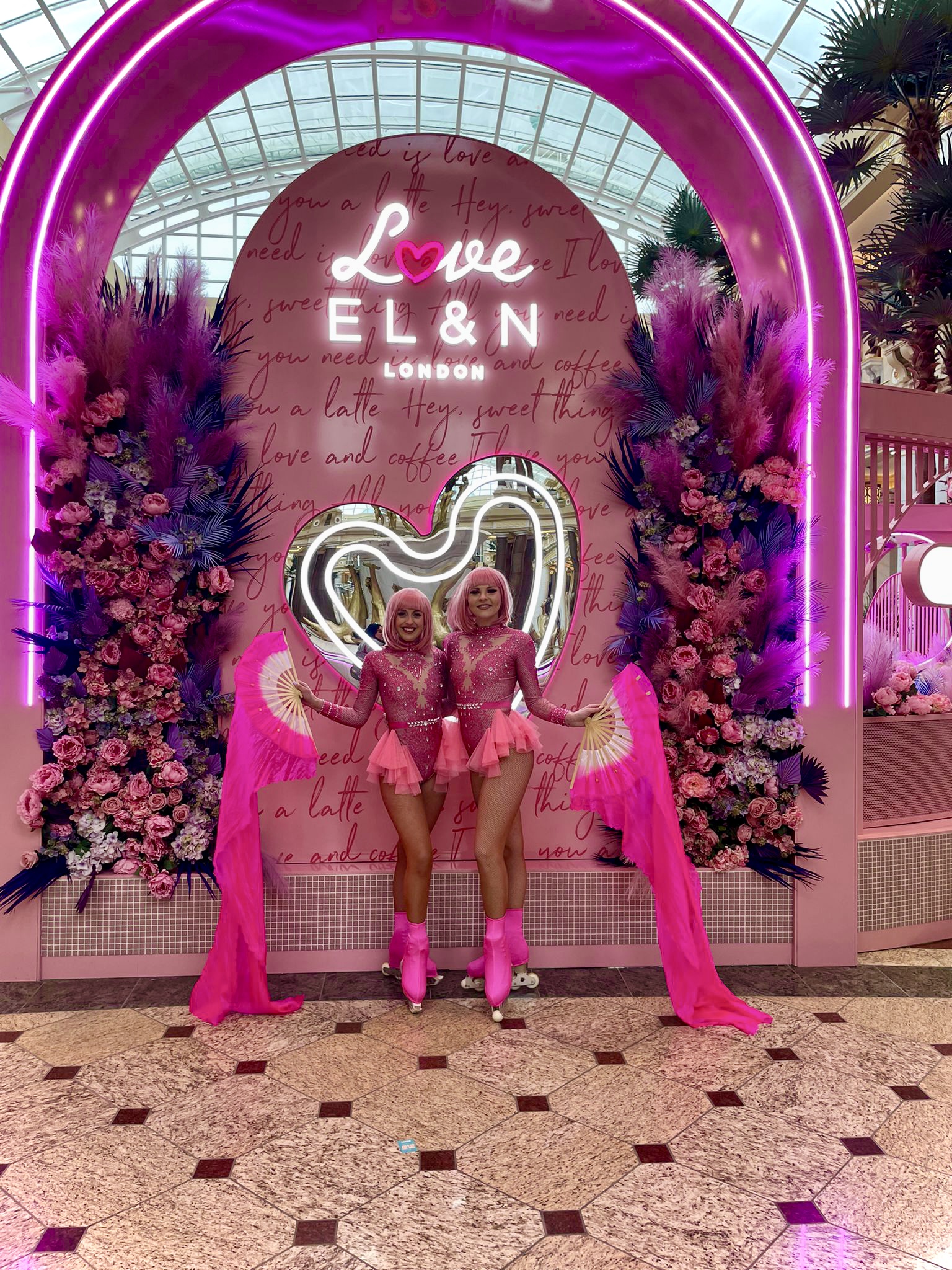 A vibrant entrance features two performers in pink outfits with roller skates, surrounded by lush floral arrangements and neon lighting, exuding a lively atmosphere.