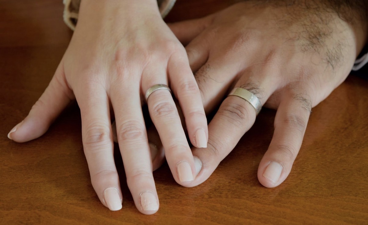 Two hands with silver wedding bands rest together on a warm wooden table, lit softly for an intimate, polished catering or event moment.