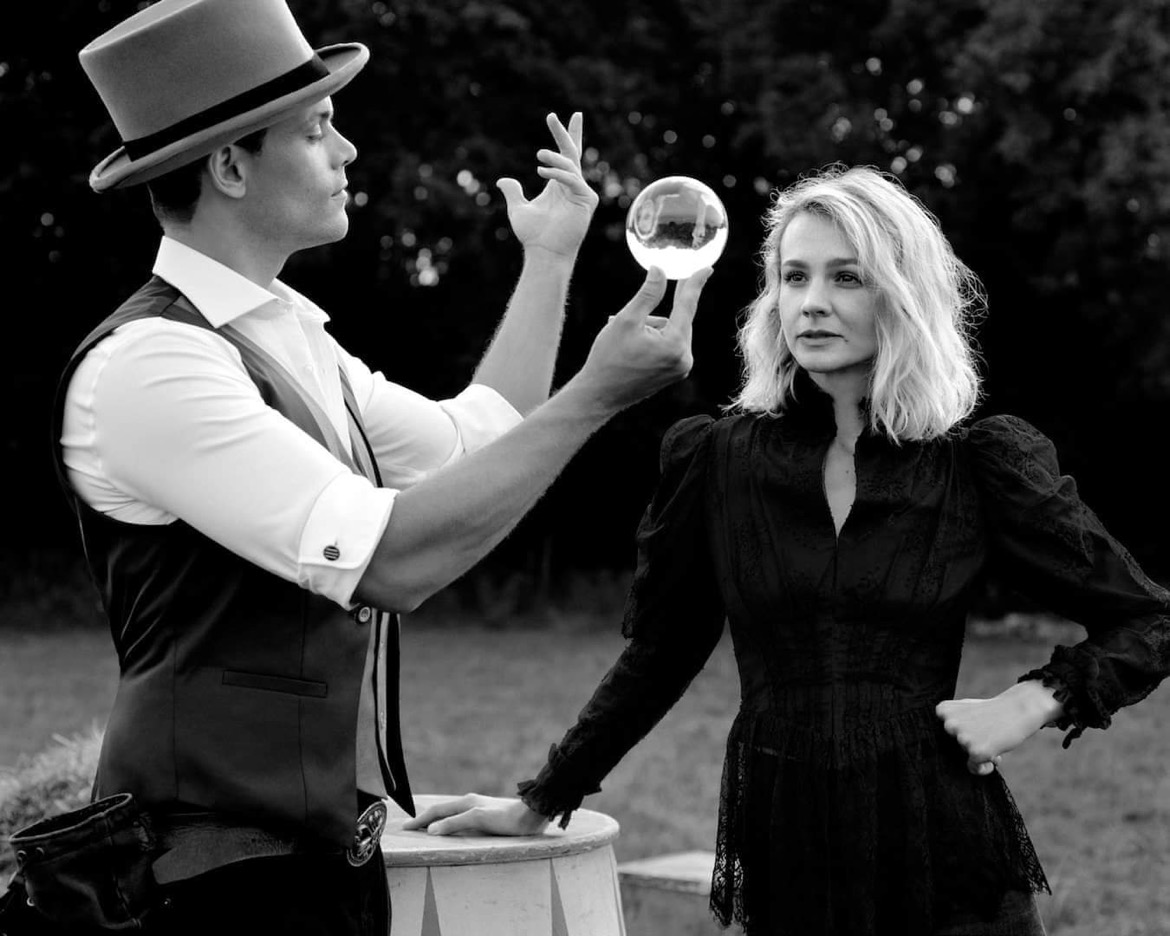 A magician in a tailored vest and hat showcases a crystal orb, while a woman in a lace black dress gazes intently, set against a moody backdrop.