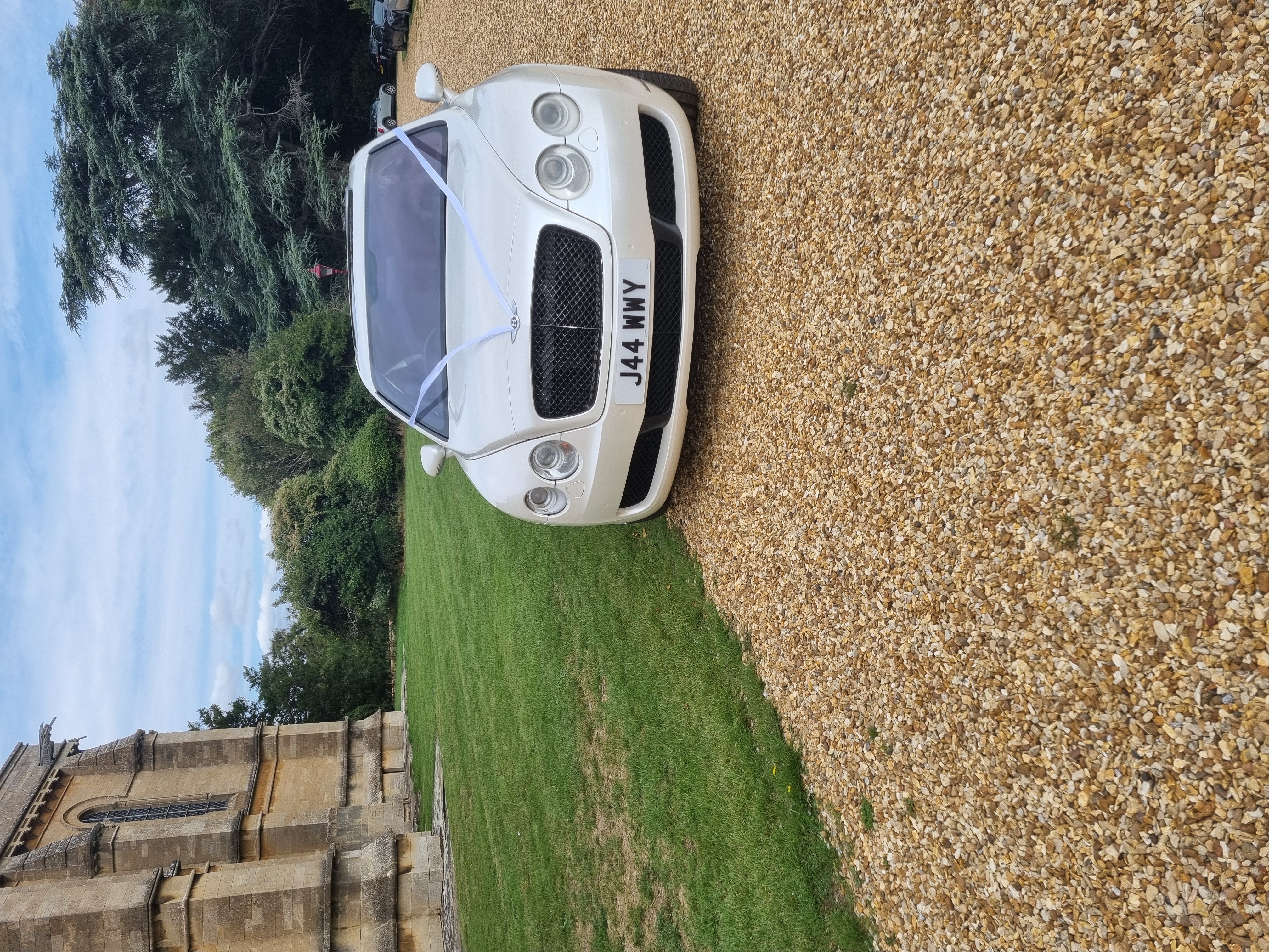 A sleek white Bentley is parked elegantly on a cobblestone drive in front of an opulent, vintage mansion under a partly cloudy sky.