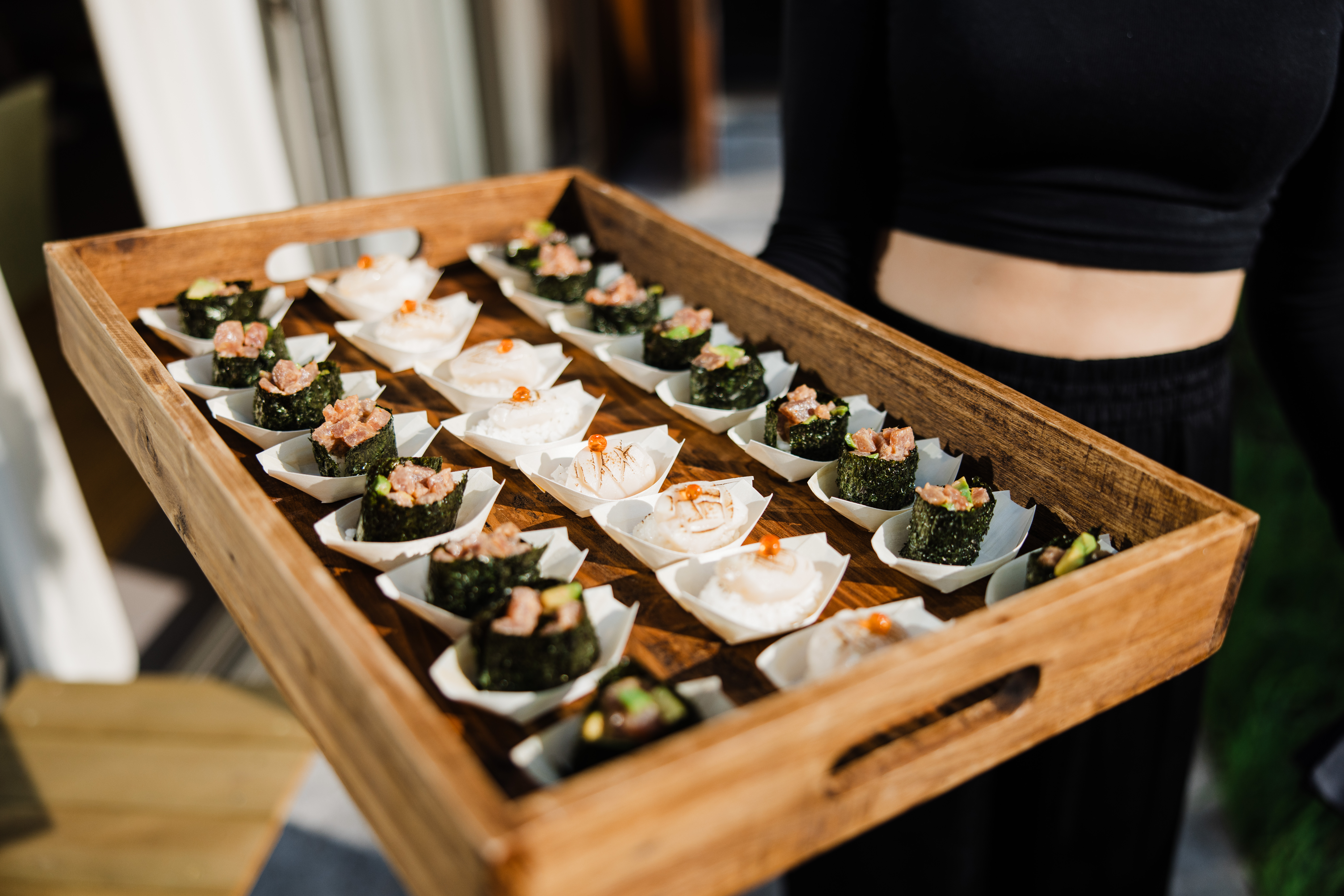 Wooden catering tray holds neatly arranged sushi bites in white paper cups, warm daylight highlighting polished, artful buffet presentation.
