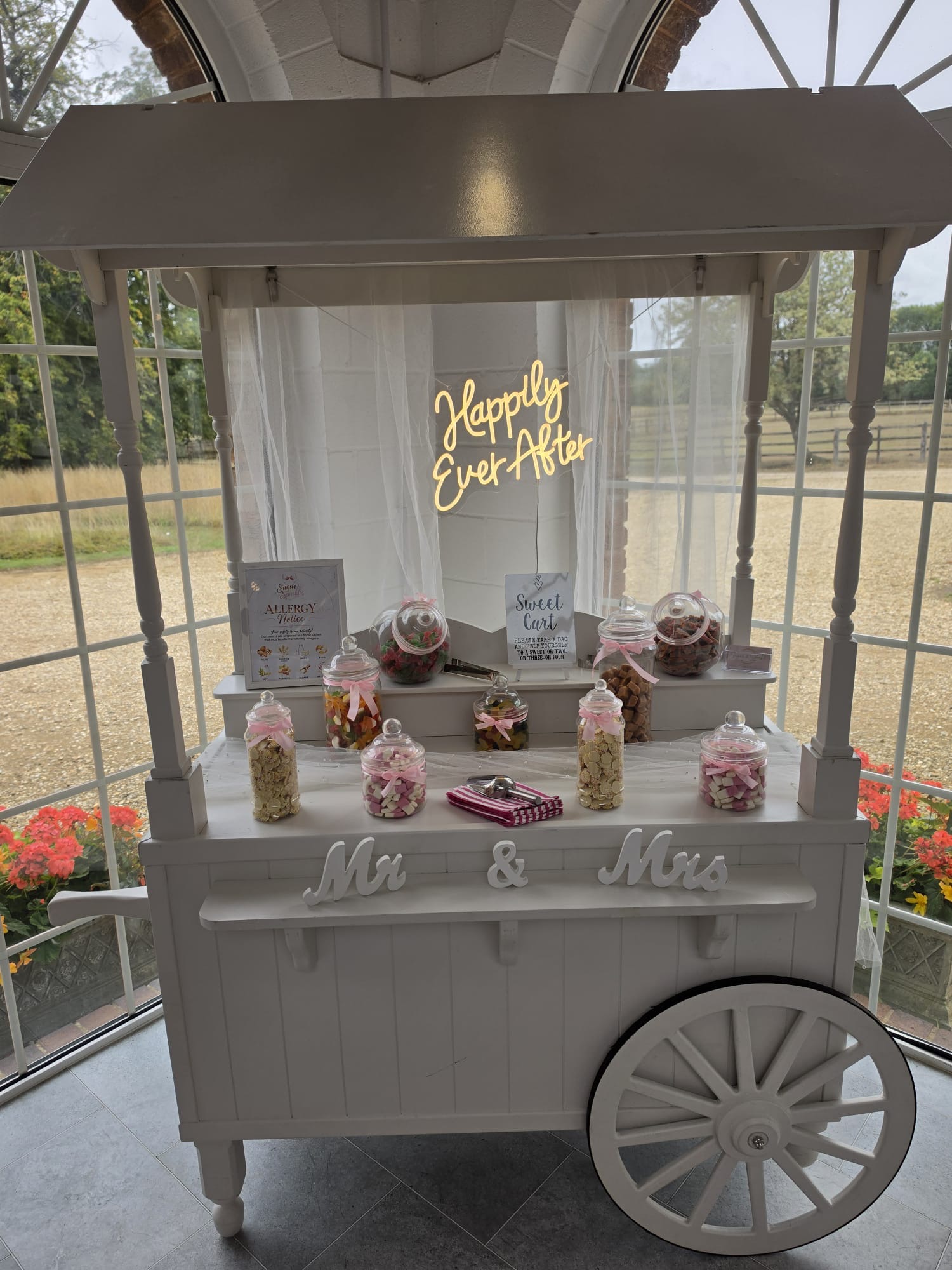 Elegant white vintage cart buffet with pink-ribbon candy jars, neon “Happily Ever After,” and crisp wedding styling in soft daylight.