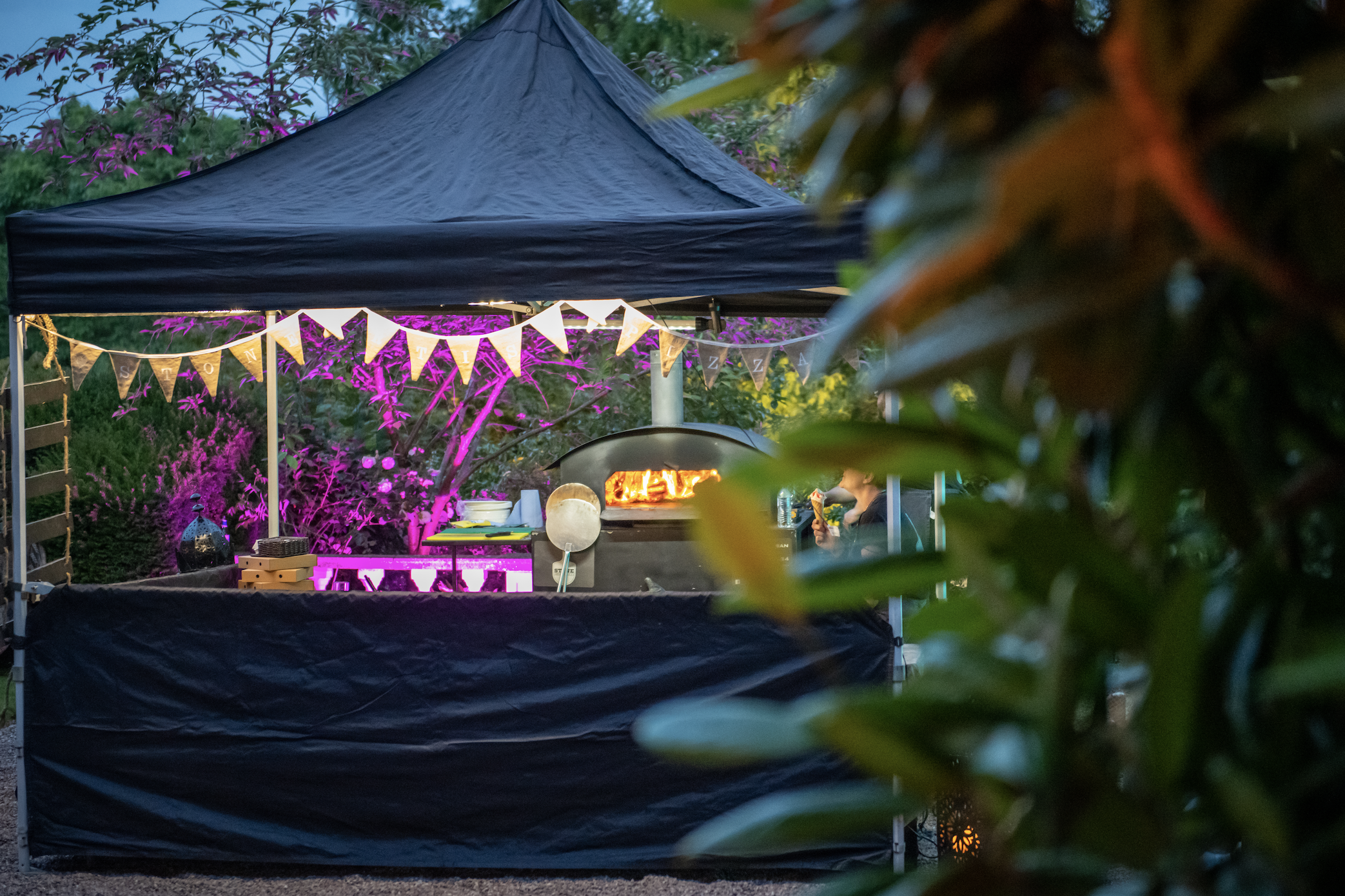 Black pop-up catering tent with glowing pizza oven, bunting, and magenta uplights, creating a polished outdoor buffet atmosphere.