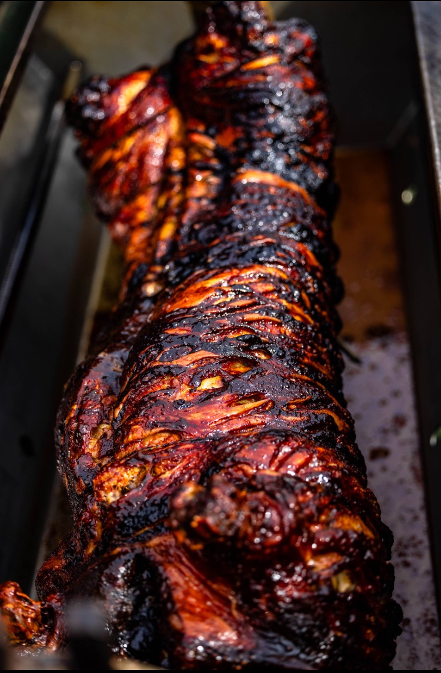 A beautifully charred whole roast, glistening in rich, dark mahogany hues, rests in a metal container, showcasing exquisite barbecue mastery.