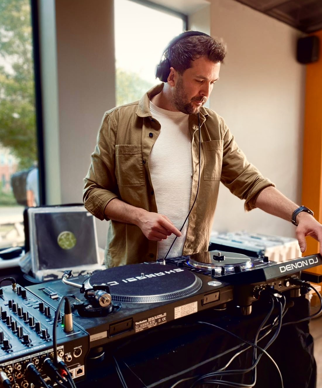Professional DJ in headphones mixing on Denon turntables and a mixer, with cables neatly spread at a bright indoor catering event.