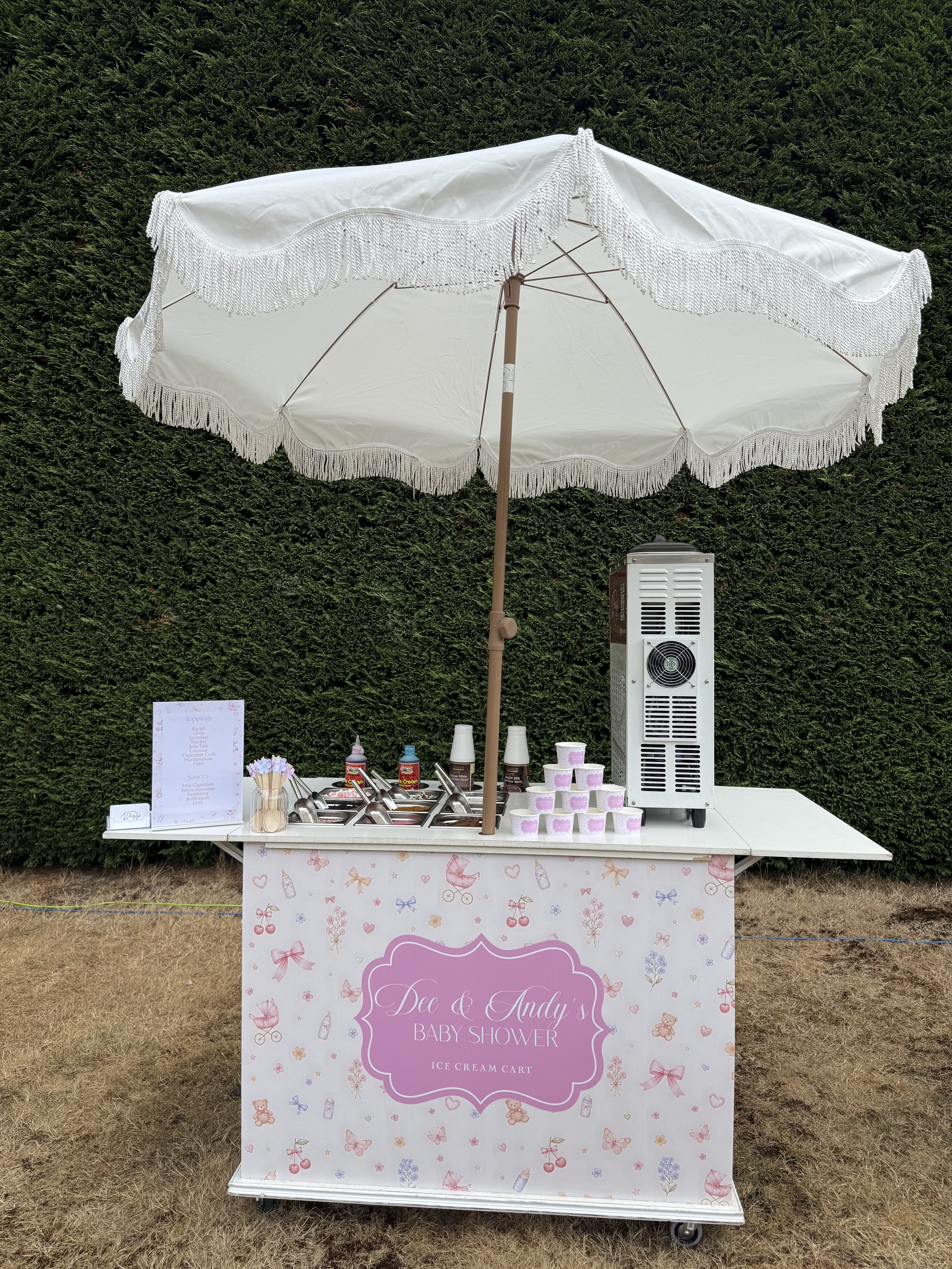 White fringed umbrella shades a pastel baby-shower ice cream cart with toppings, pink cups, and a polished freezer against a hedge.