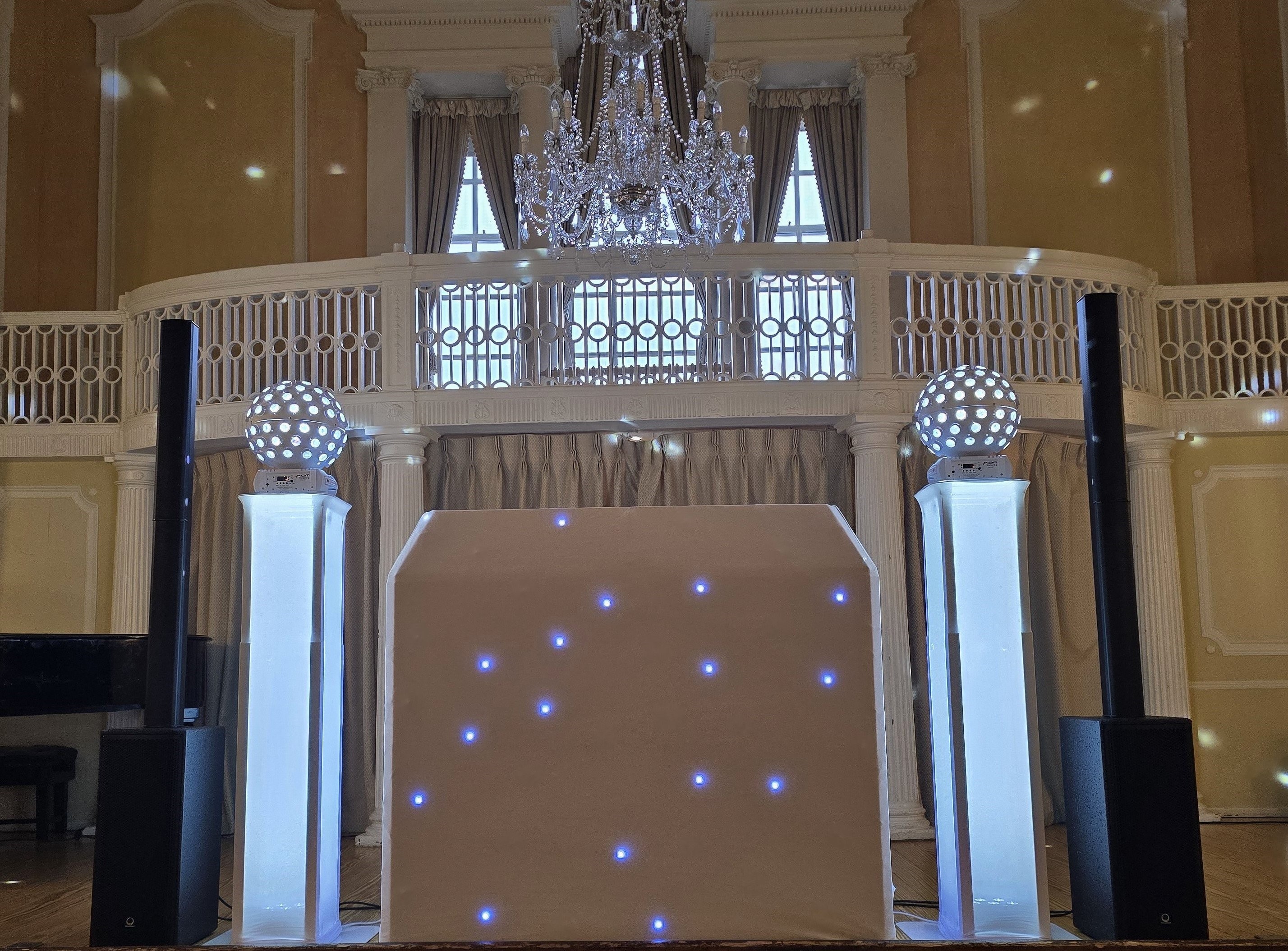 Elegant wedding DJ setup under a grand chandelier, with glowing LED pillars and disco spheres framing a starry white booth in warm ballroom light.