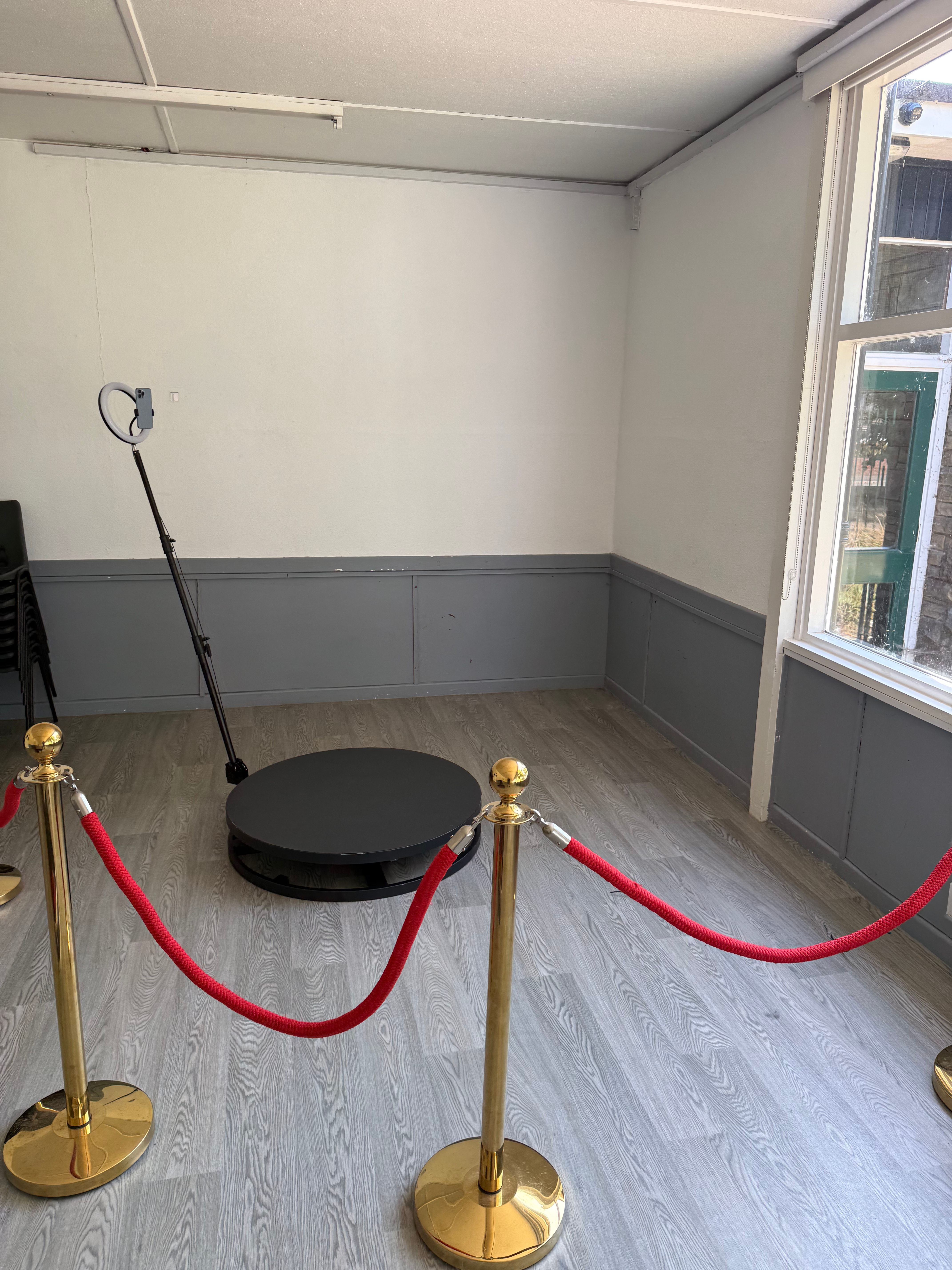 Empty event room with ring light and black platform, roped off by red velvet stanchions, bright daylight, polished service setup.
