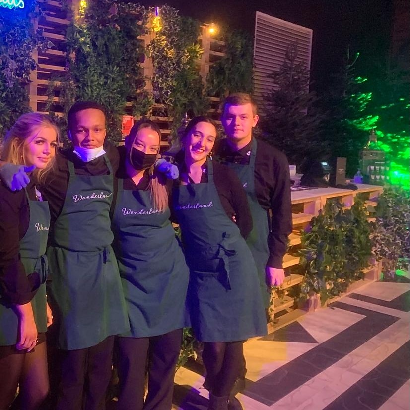 Smiling catering staff in matching Wonderland aprons pose beside a leafy outdoor buffet, under moody purple-green event lighting.