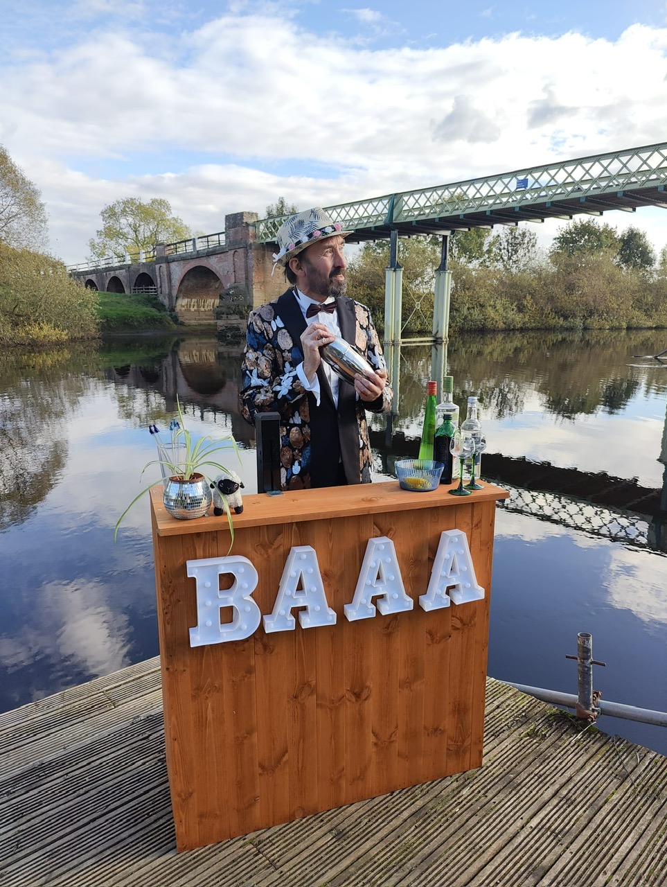 A dapper bartender in a floral suit mixes drinks at a wooden bar beside a serene river, reflecting vibrant colors under bright daylight.