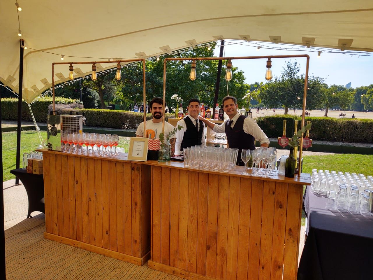 A rustic wooden bar displays elegant glassware and colorful cocktails, staffed by smiling attendants, under bright daylight in a charming park.