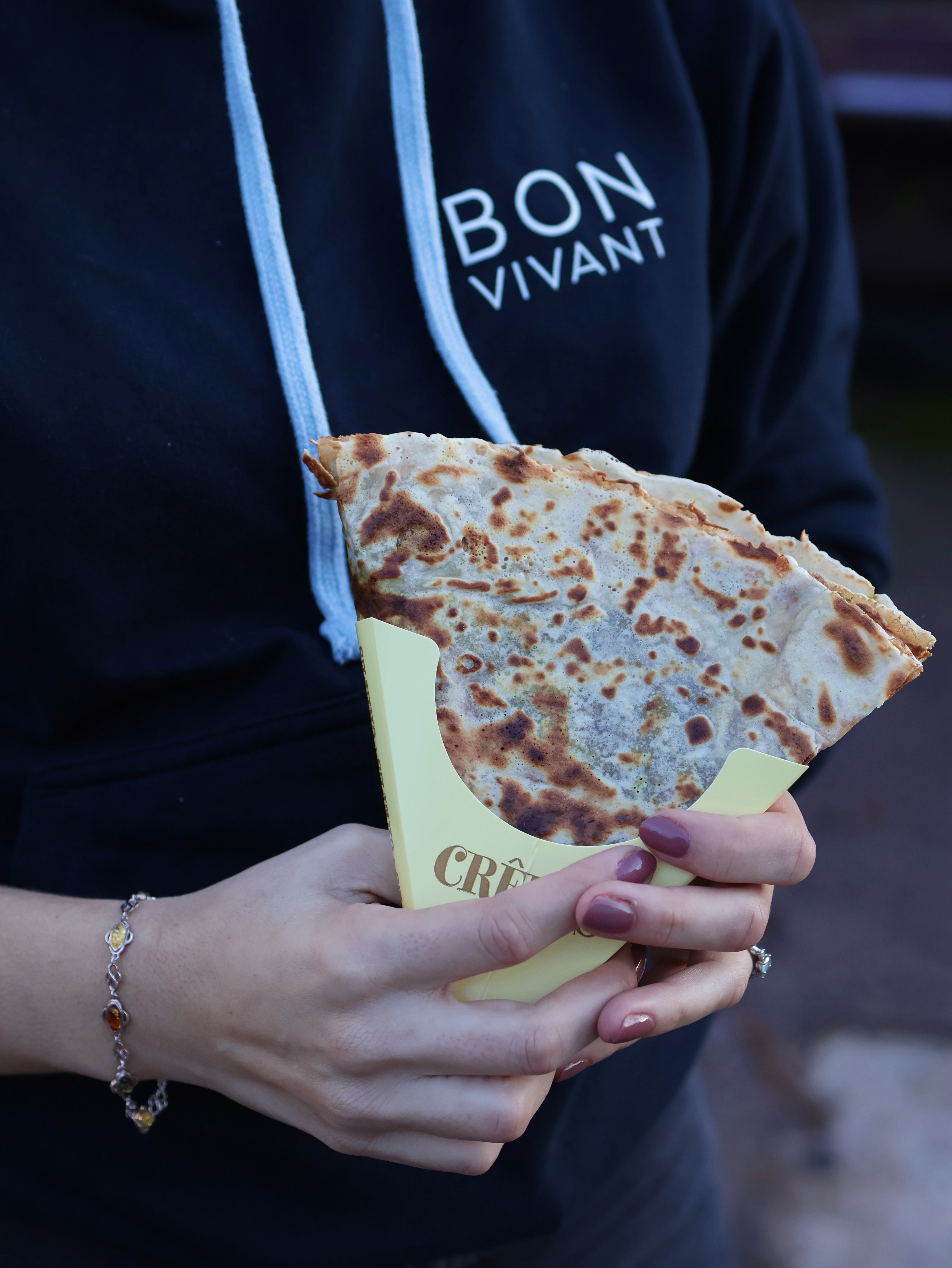 A hand holds a golden-brown crepe in a yellow paper holder against a dark hoodie, showcasing casual street food vibes perfect for catering.