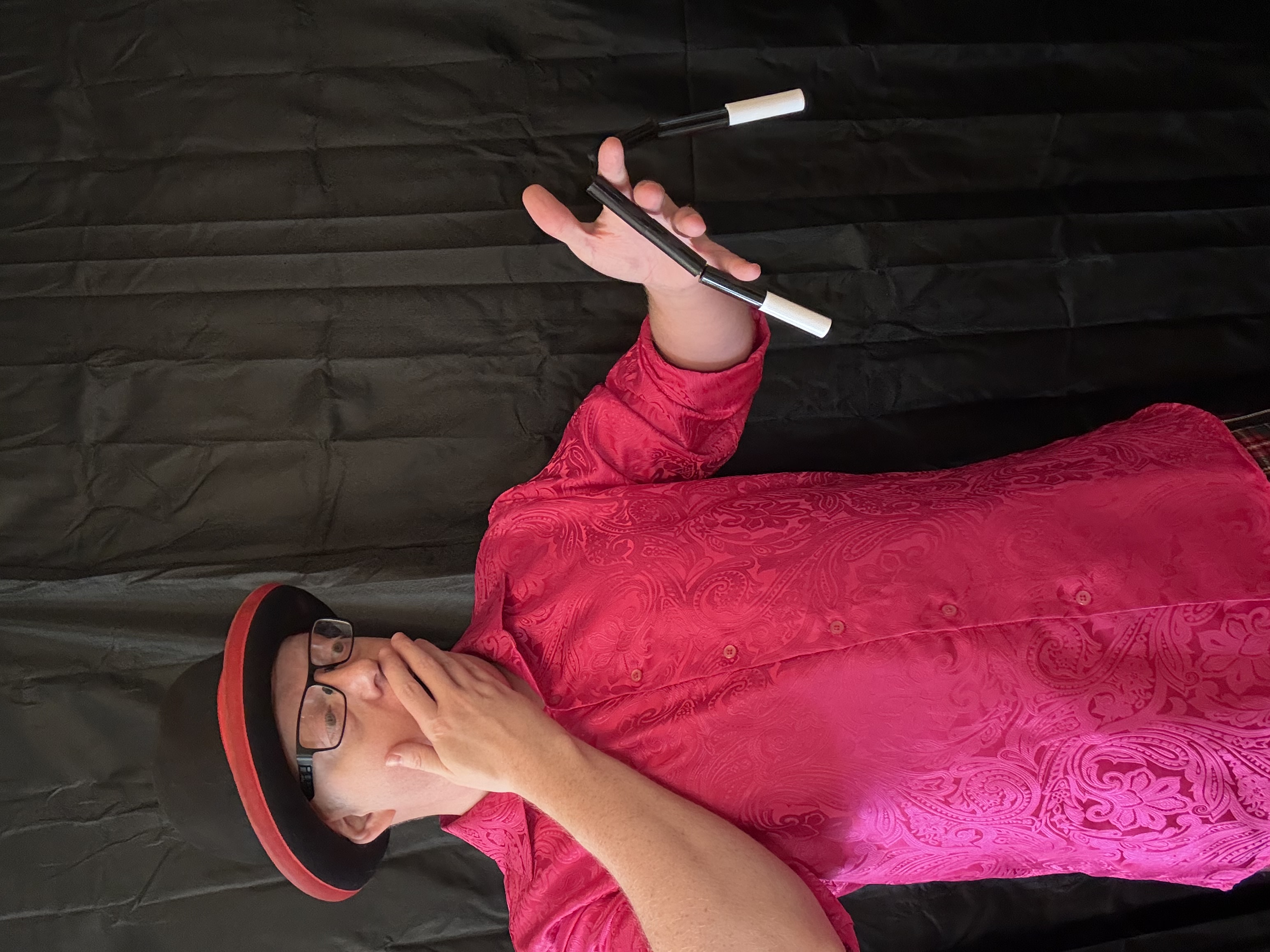 A magician in a vibrant pink patterned shirt and top hat performs an astonished act with black-and-white props against a dark backdrop.