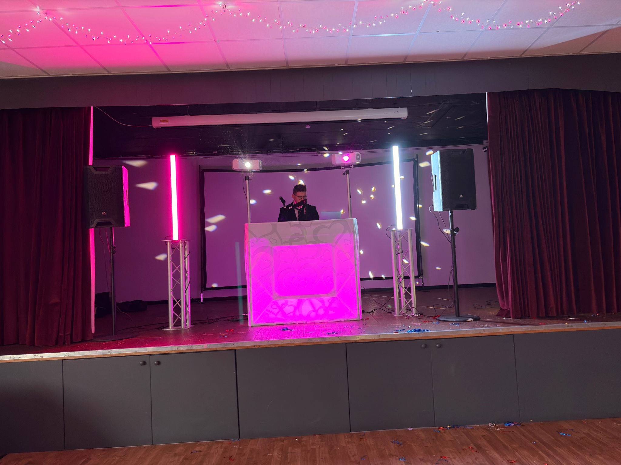 Pink-lit DJ booth on a small stage, flanked by speakers and LED columns, with disco reflections and a polished event venue vibe.