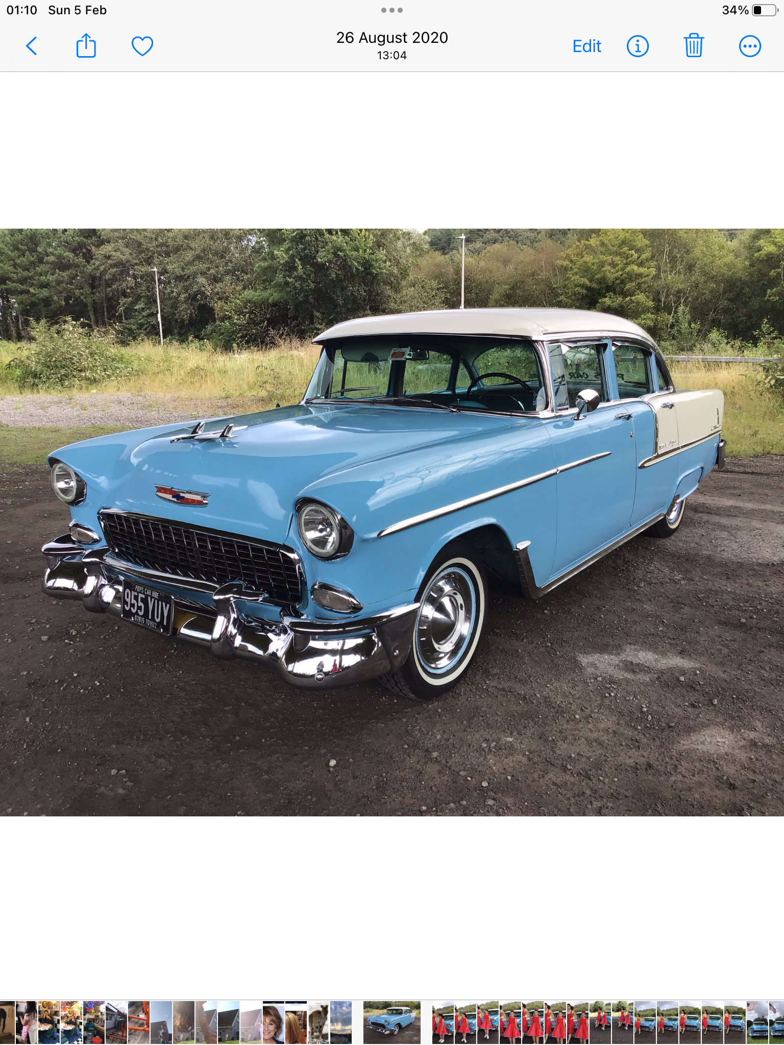 A vintage blue and white classic car shines under soft daylight, parked on gravel with lush greenery as a backdrop, showcasing timeless elegance.