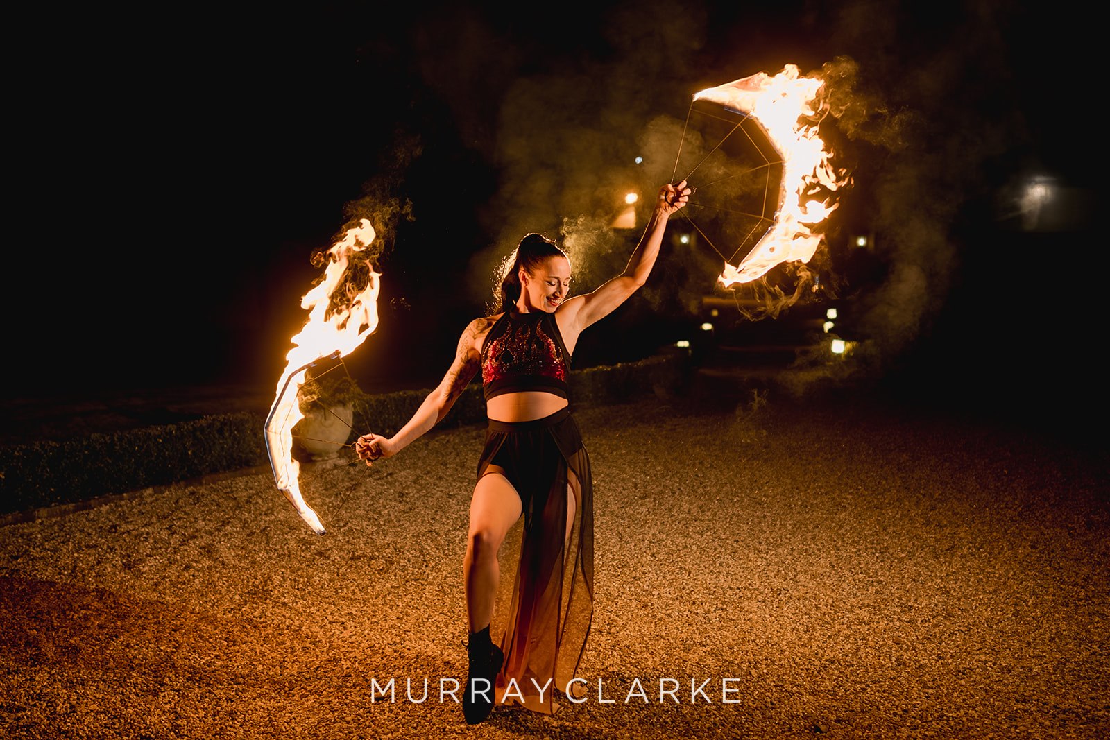 A fire performer dances energetically at night, wielding flaming torches against a dark backdrop, creating a vibrant, thrilling atmosphere.