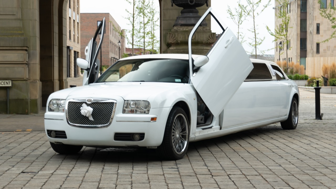 White stretch limousine with both rear doors open, parked on cobblestones in a city plaza, evoking polished wedding transport elegance.