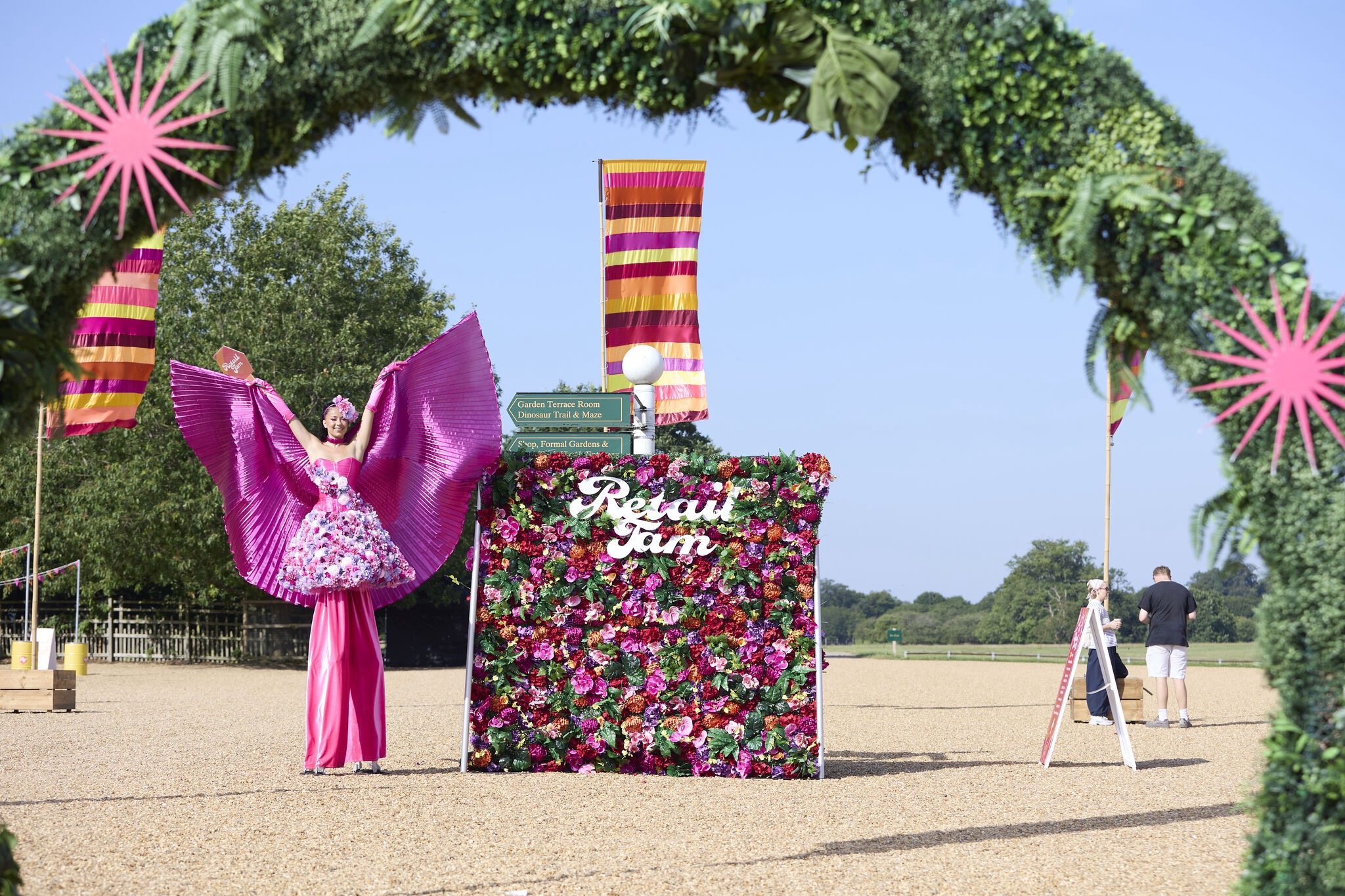 Bright outdoor festival display with a pink-wing performer, floral photo wall, striped banners, and lush greenery, under clear daylight.