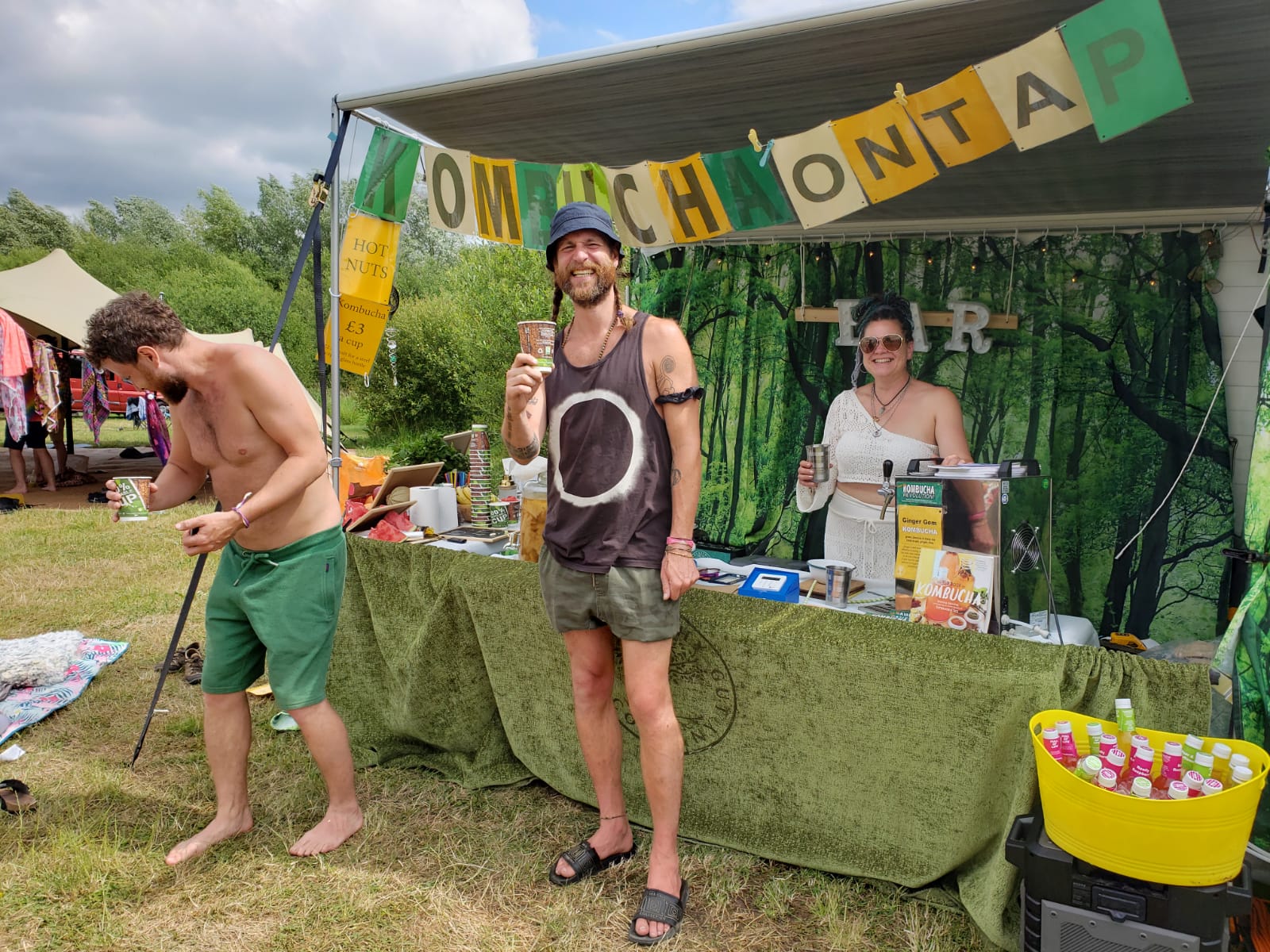A vibrant outdoor kombucha bar features friendly staff, colorful cups, and fresh drinks displayed on a green cloth. Sunlit, festival vibes.