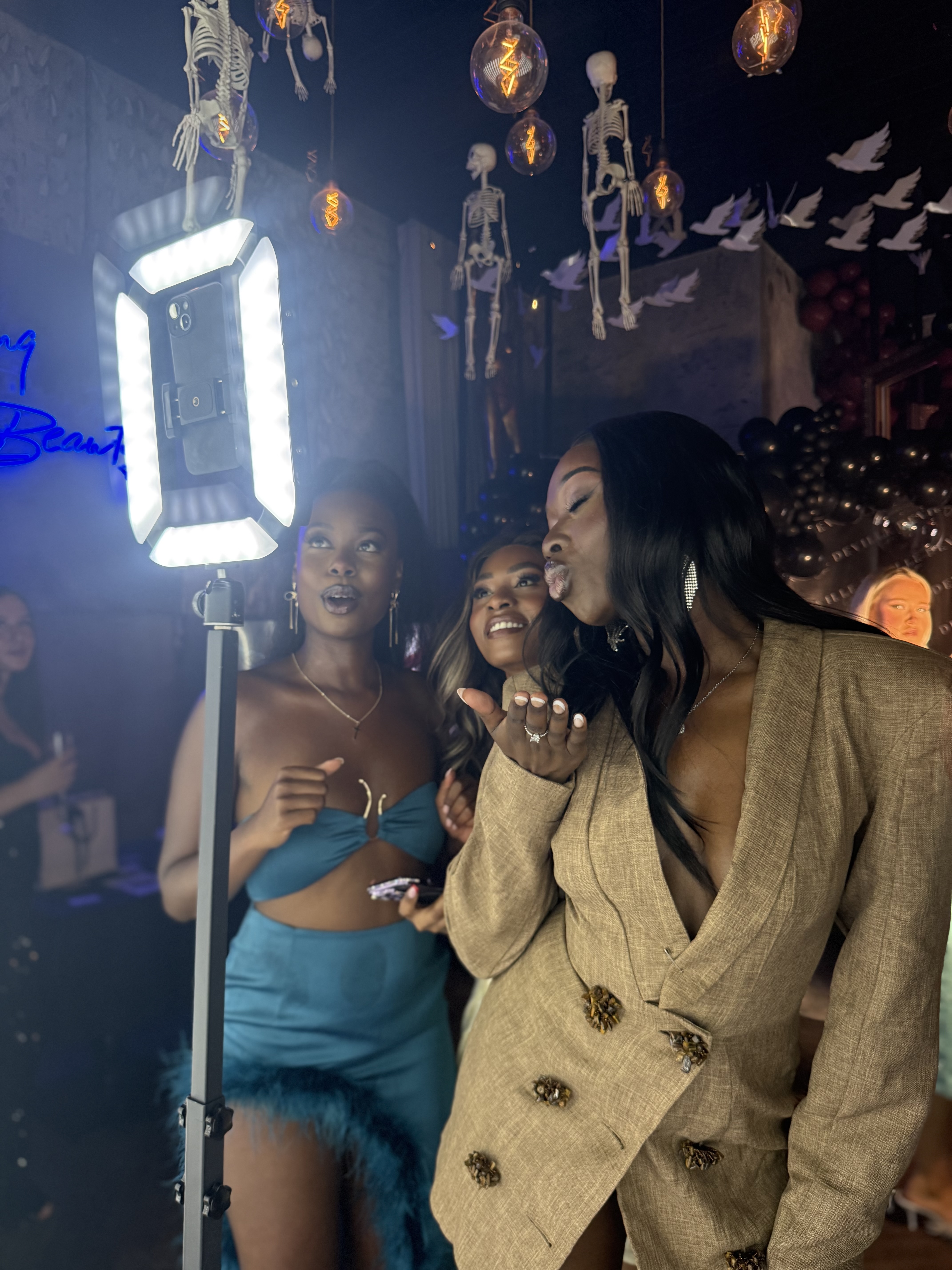Three women pose playfully near a ring light in a lively venue, adorned with vintage skeleton decorations and glowing pendant lights.