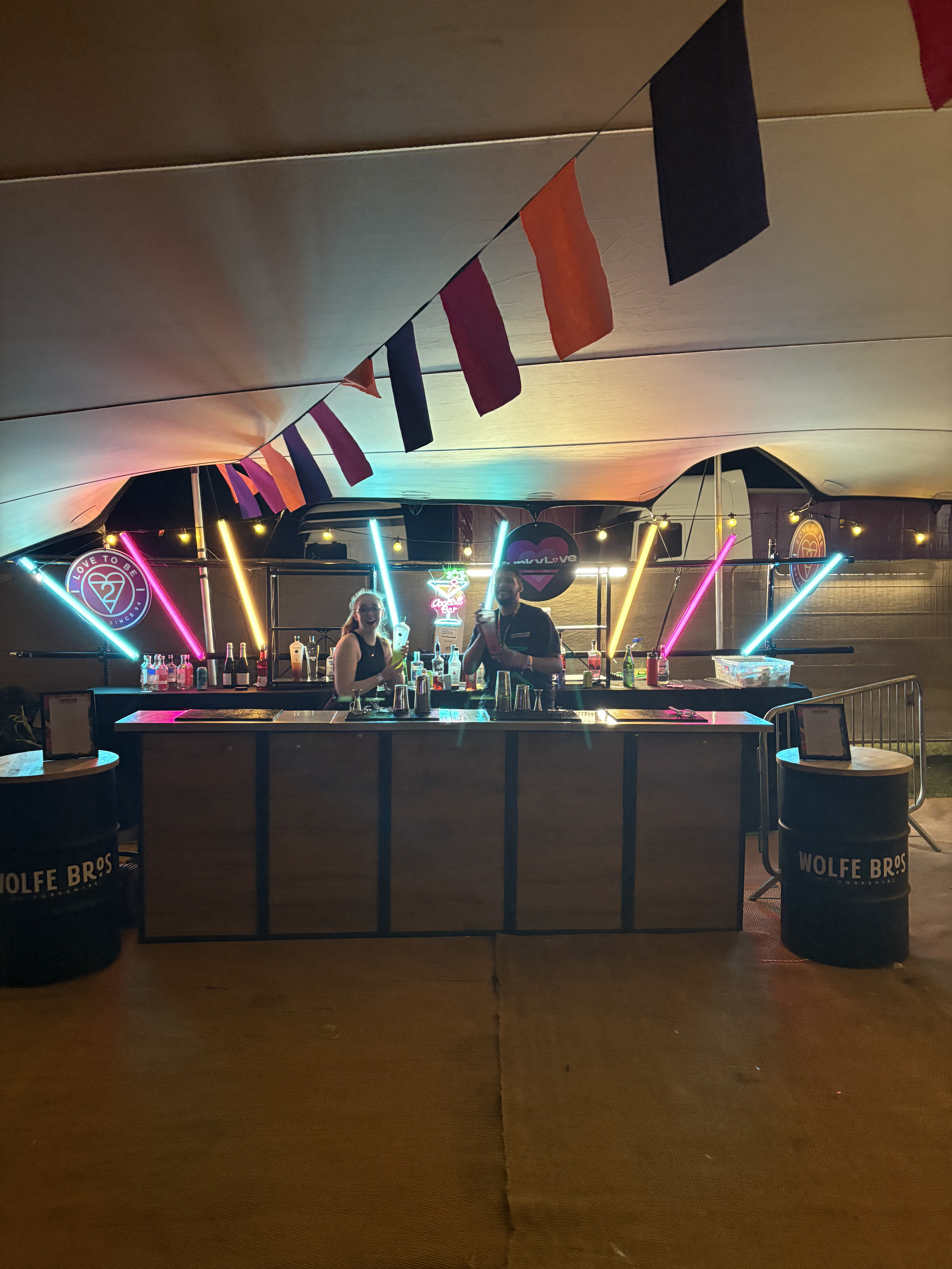 A vibrant outdoor bar features neon lighting and colorful bunting, with two bartenders mixing drinks at a sleek wooden counter, enhancing the festive atmosphere.