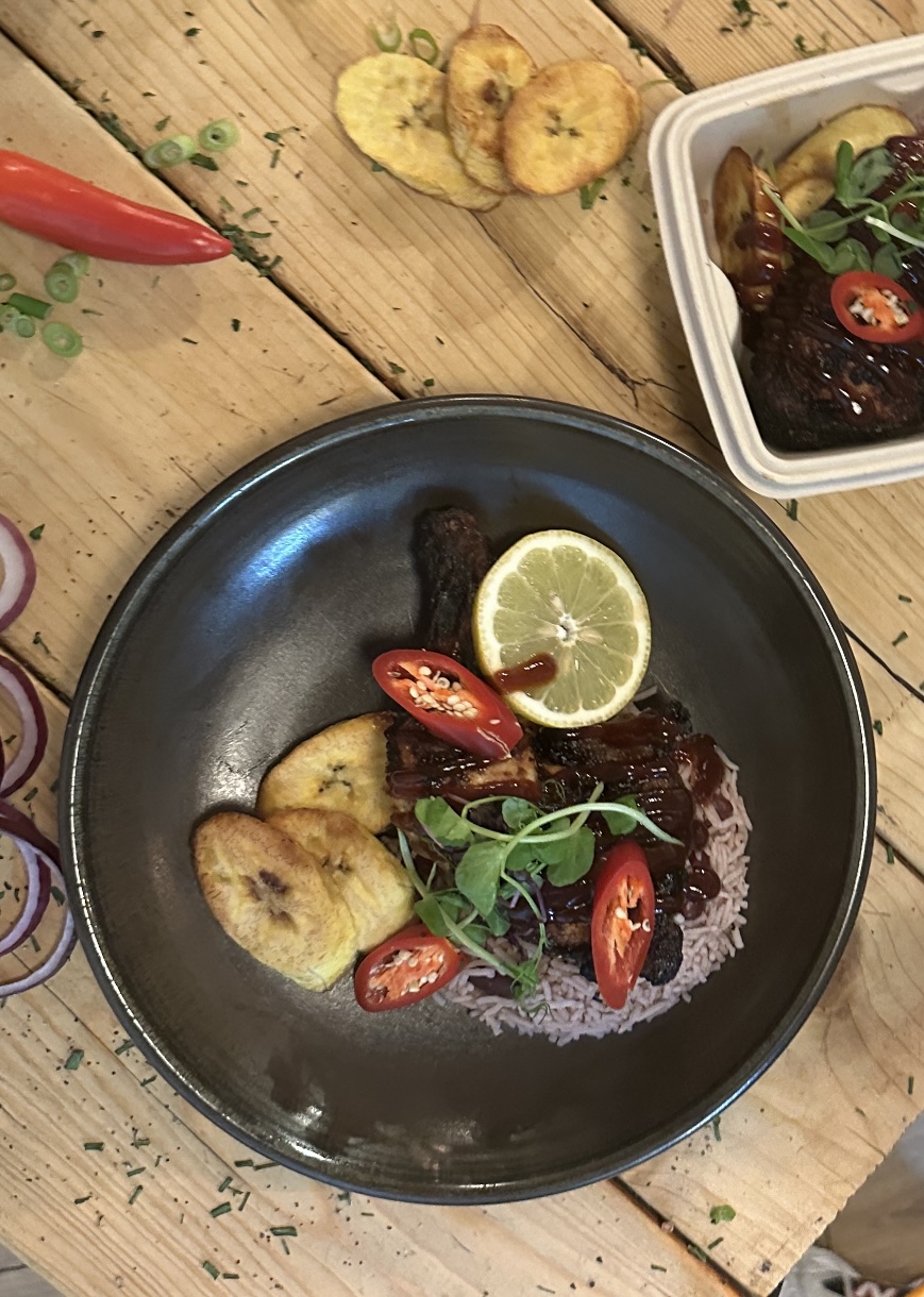 A dark plate features succulent meat, ripe lime, fresh herbs, and vibrant red chilies beside fried plantains, on rustic wood.