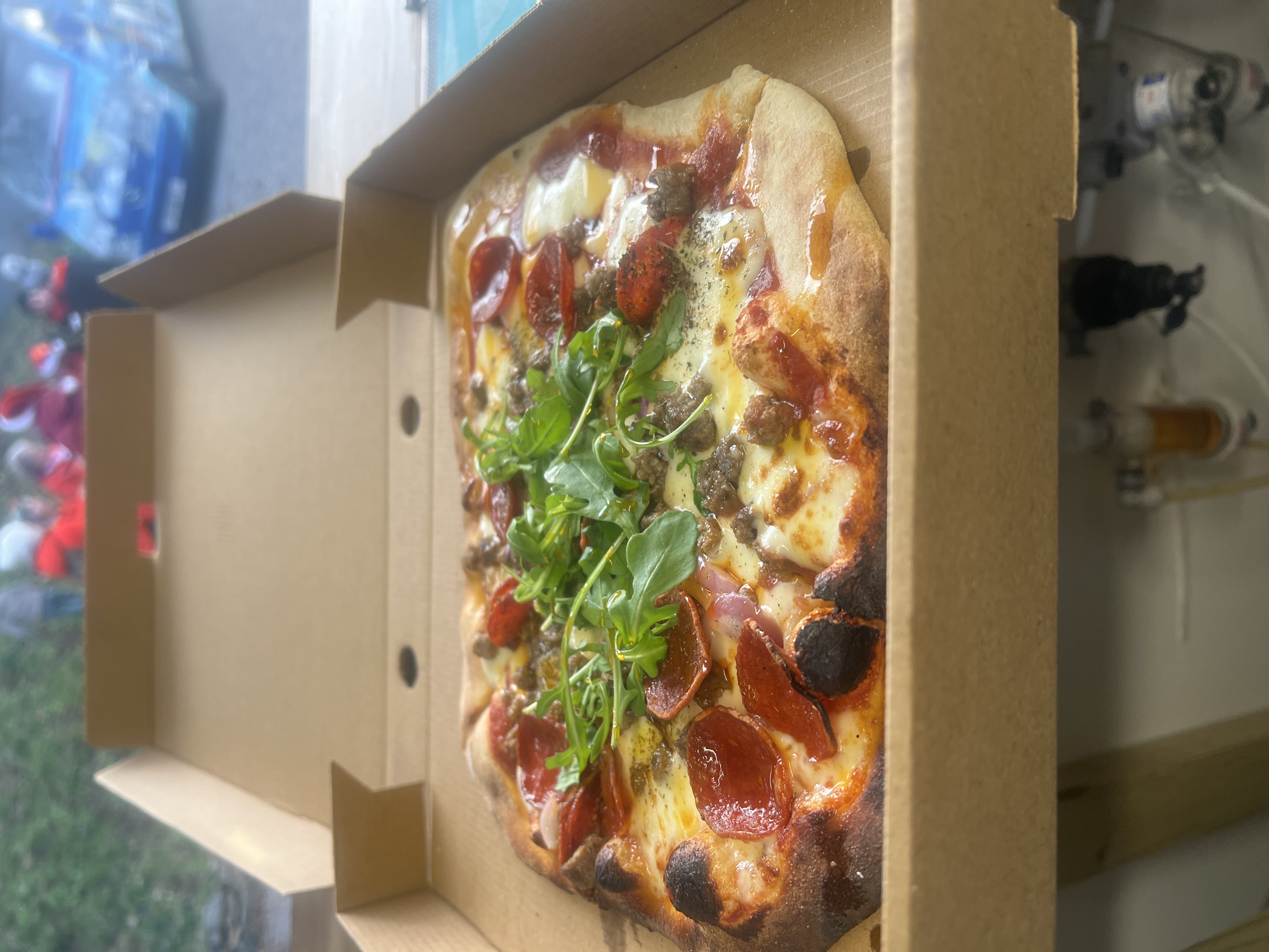 A golden-brown crust pizza topped with pepperoni, sausage, and fresh arugula sits in a cardboard box, ready for enjoyment.