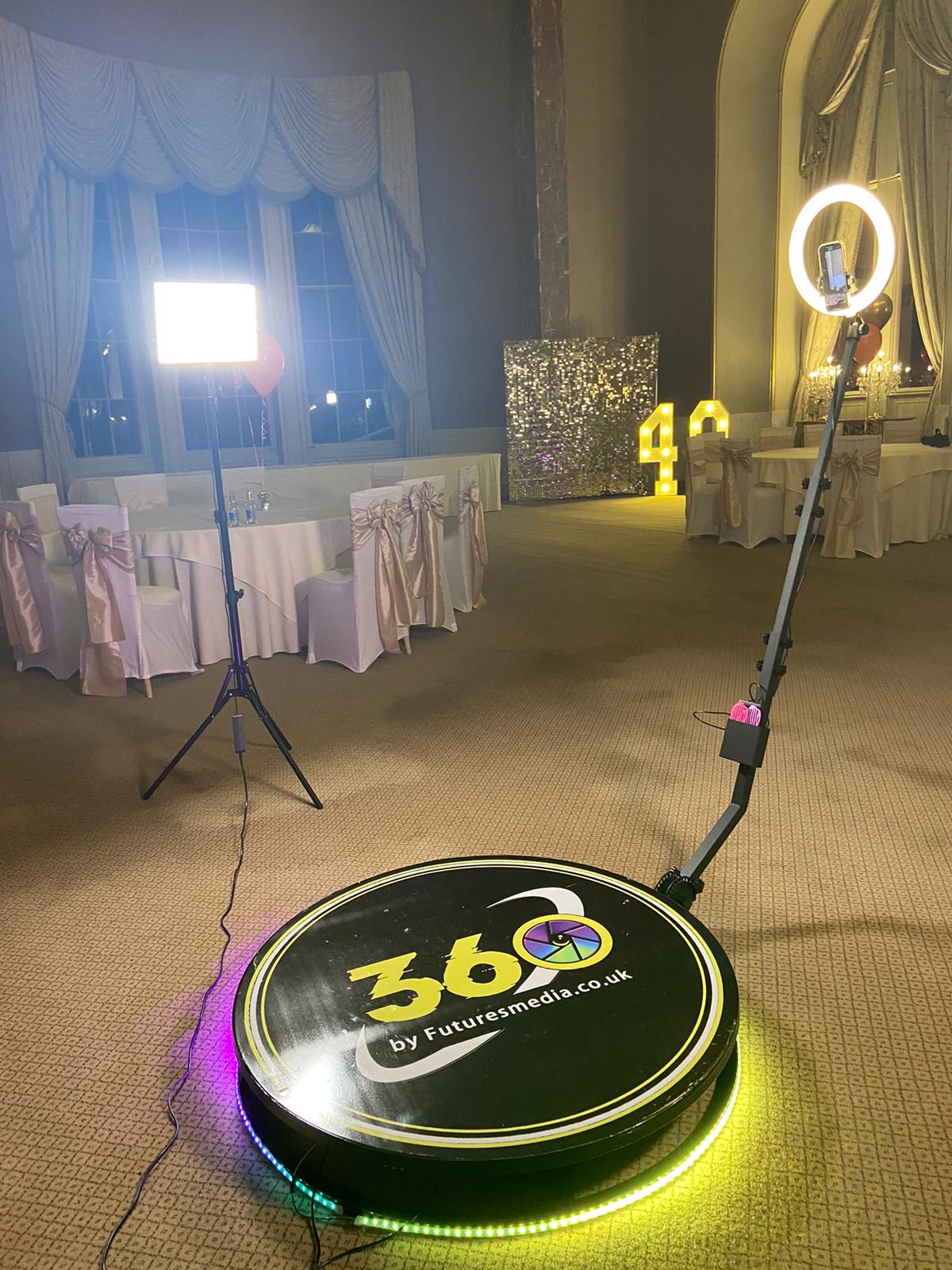 A luxurious event space features a polished 360 photo booth with vibrant LED accents, surrounded by elegantly draped chairs under warm lighting.