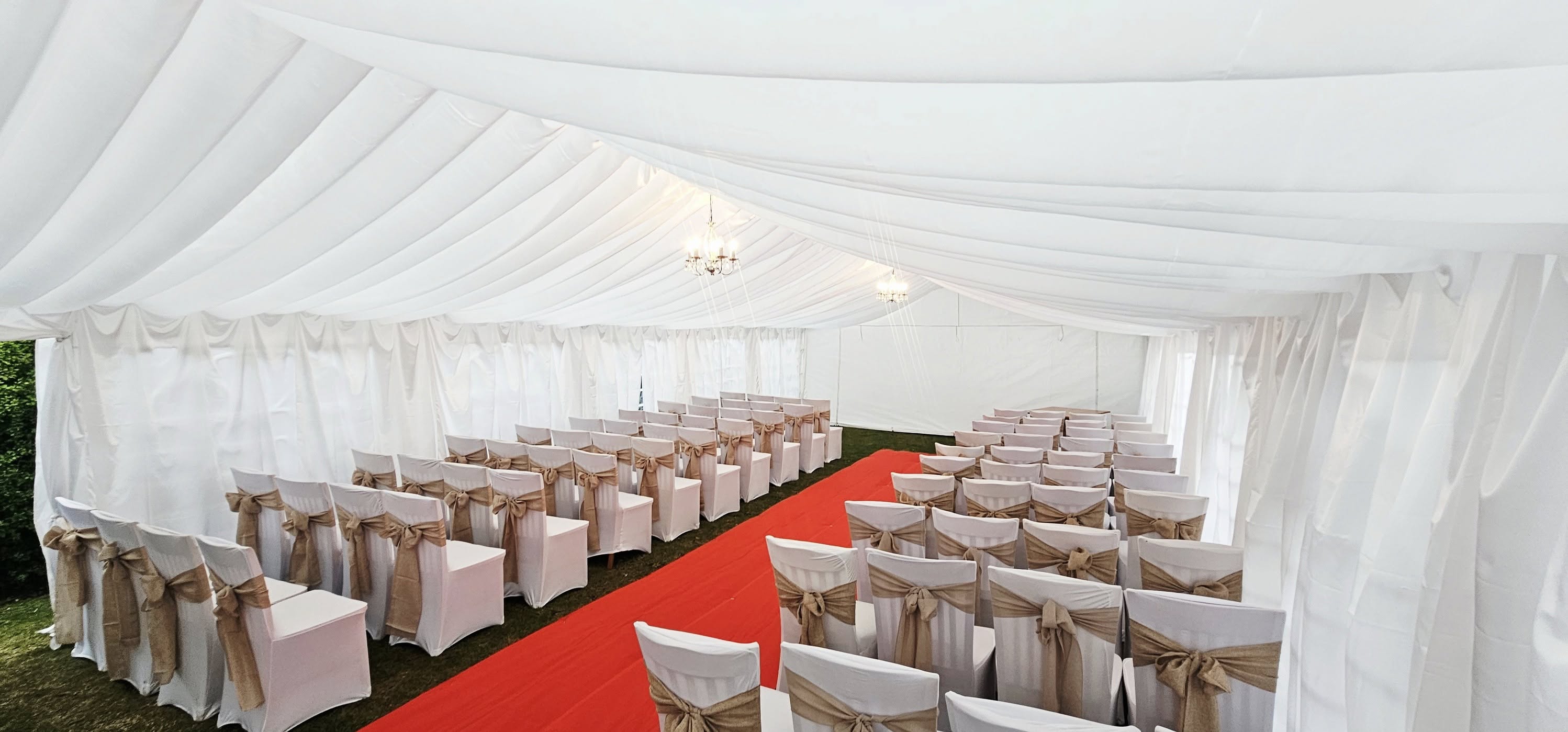 A white tent features rows of elegantly draped chairs with tan sashes, centered around a vibrant red carpet, illuminated by soft lighting.