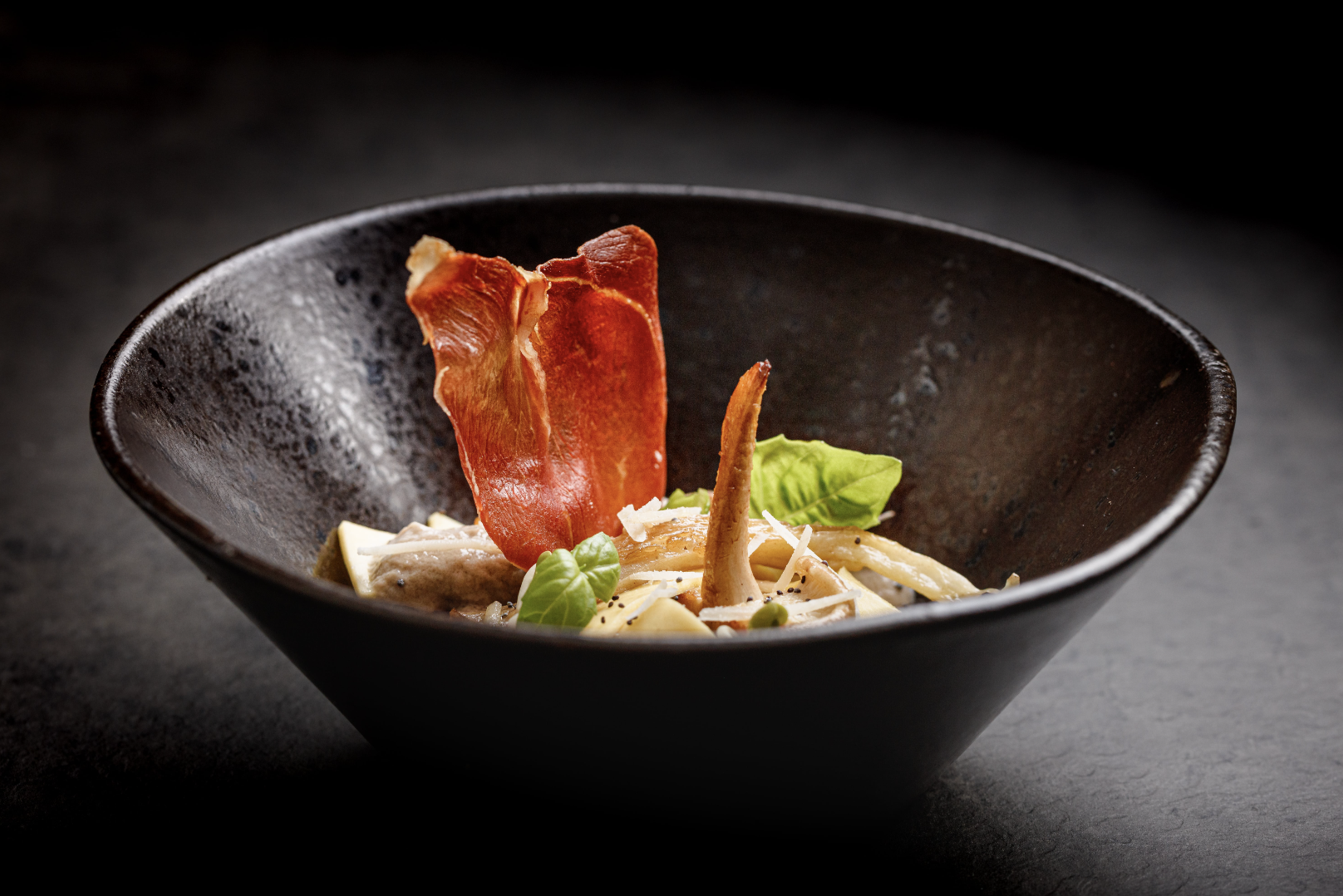 A dark, rustic bowl showcases creamy pasta topped with vibrant basil, crispy prosciutto, and earthy mushrooms, evoking elegance and quality.