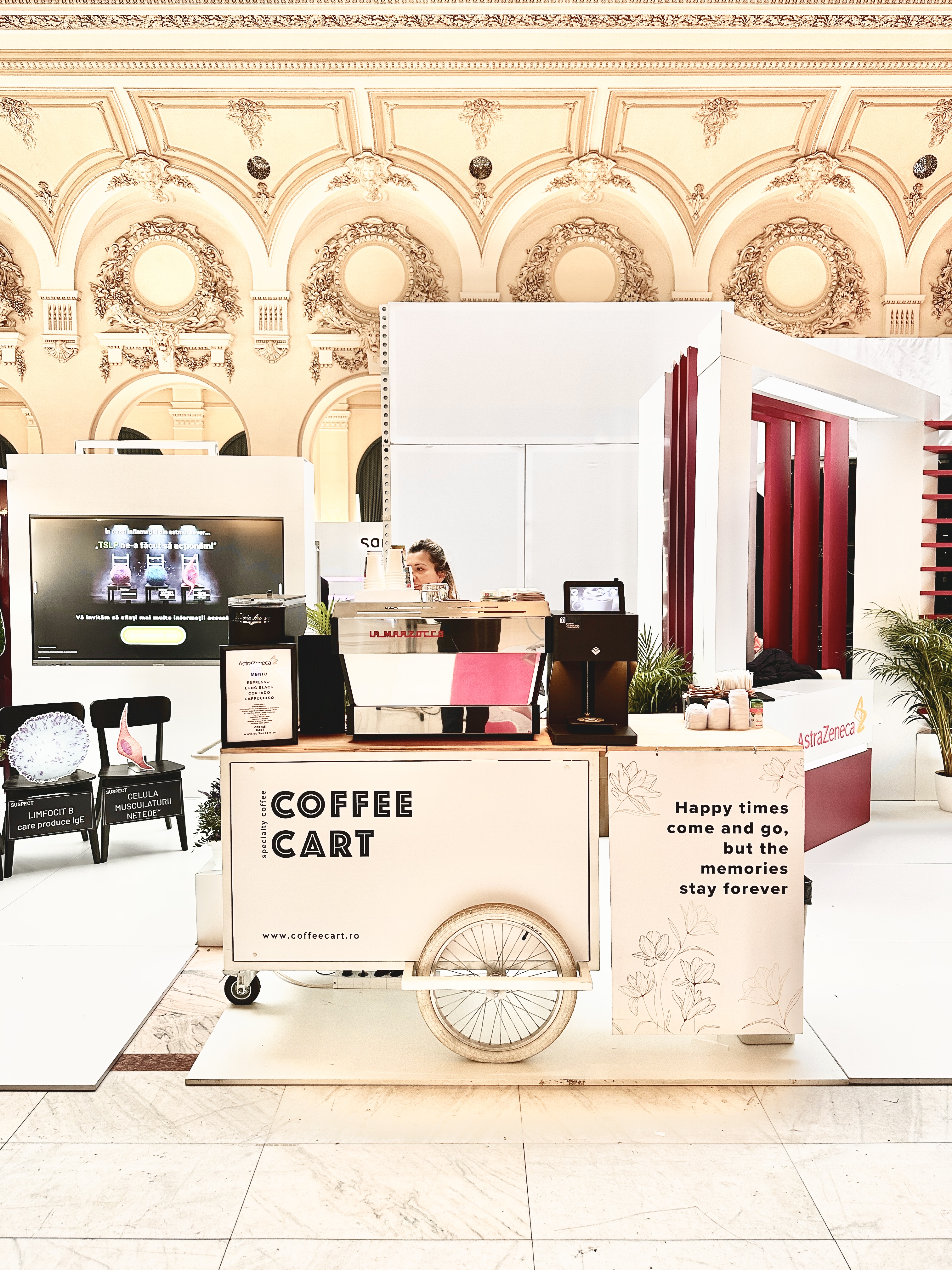 A stylish coffee cart features polished wood and a bicycle wheel, set against elegant, high-ceilinged architecture, radiating a vibrant atmosphere.