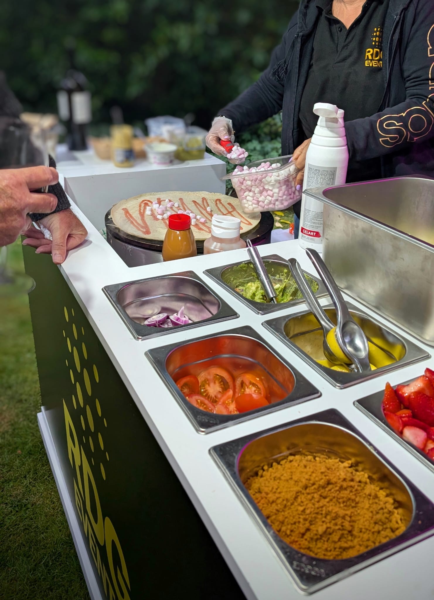 A vibrant outdoor catering setup features fresh ingredients like tomatoes, guacamole, and colorful marshmallows, presented in polished stainless trays.