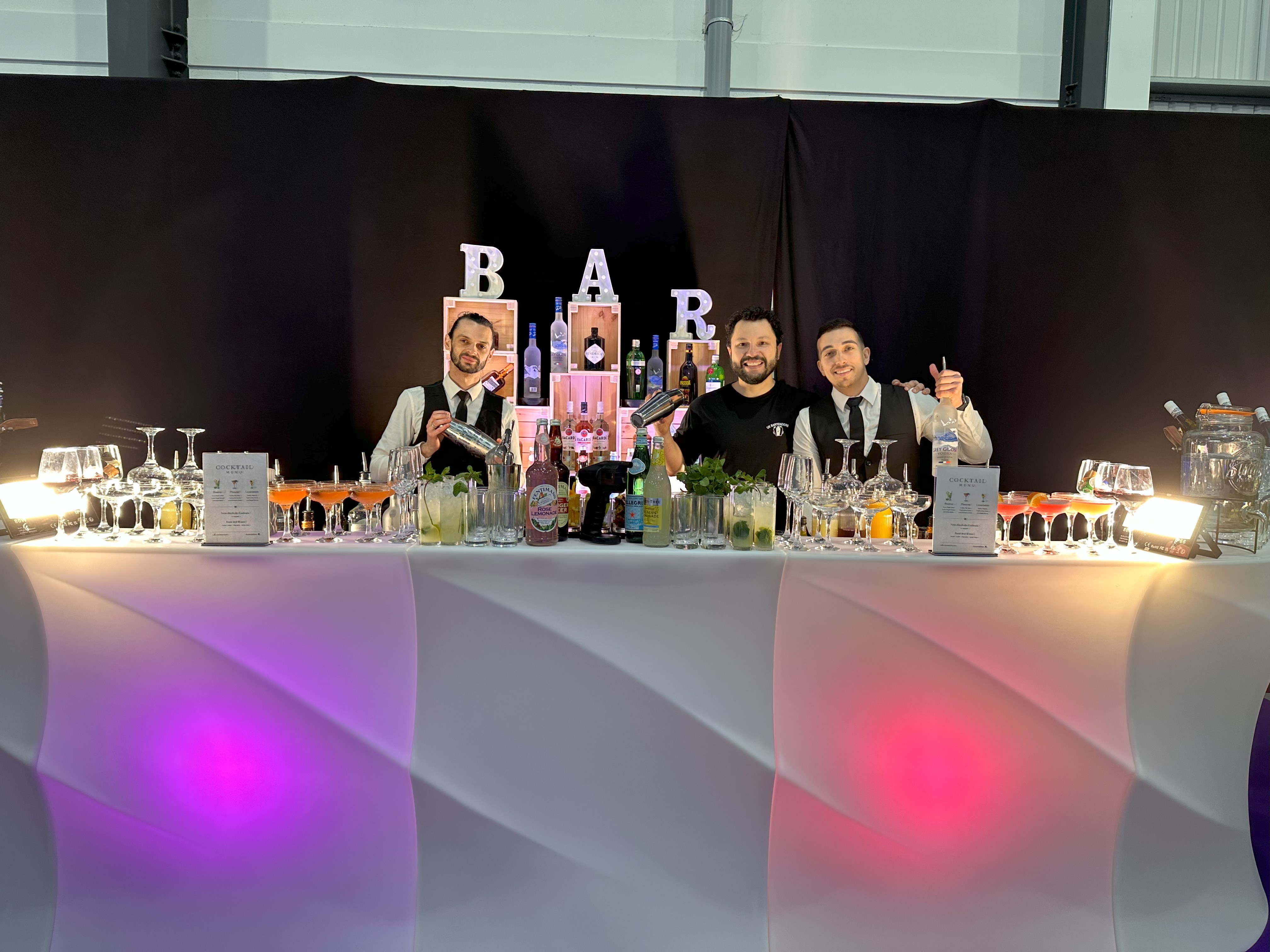 Three bartenders stand behind a sleek white bar with colorful cocktails, polished glassware, and a vibrant illuminated backdrop, exuding a festive vibe.