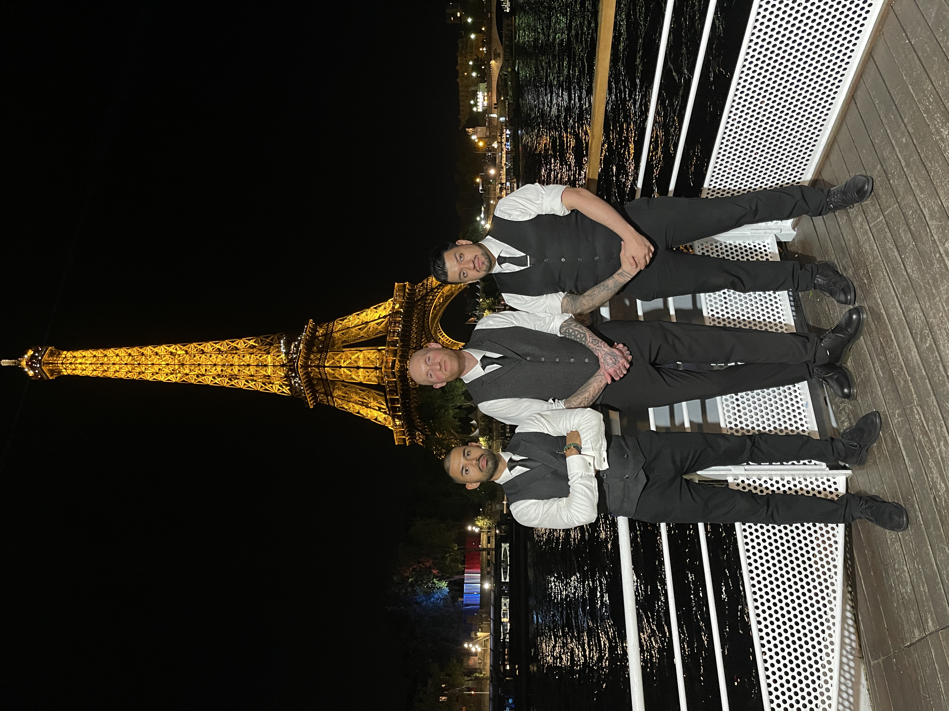 Under the romantic glow of the Eiffel Tower, three elegantly dressed men in tailored vests pose confidently against a shimmering backdrop.