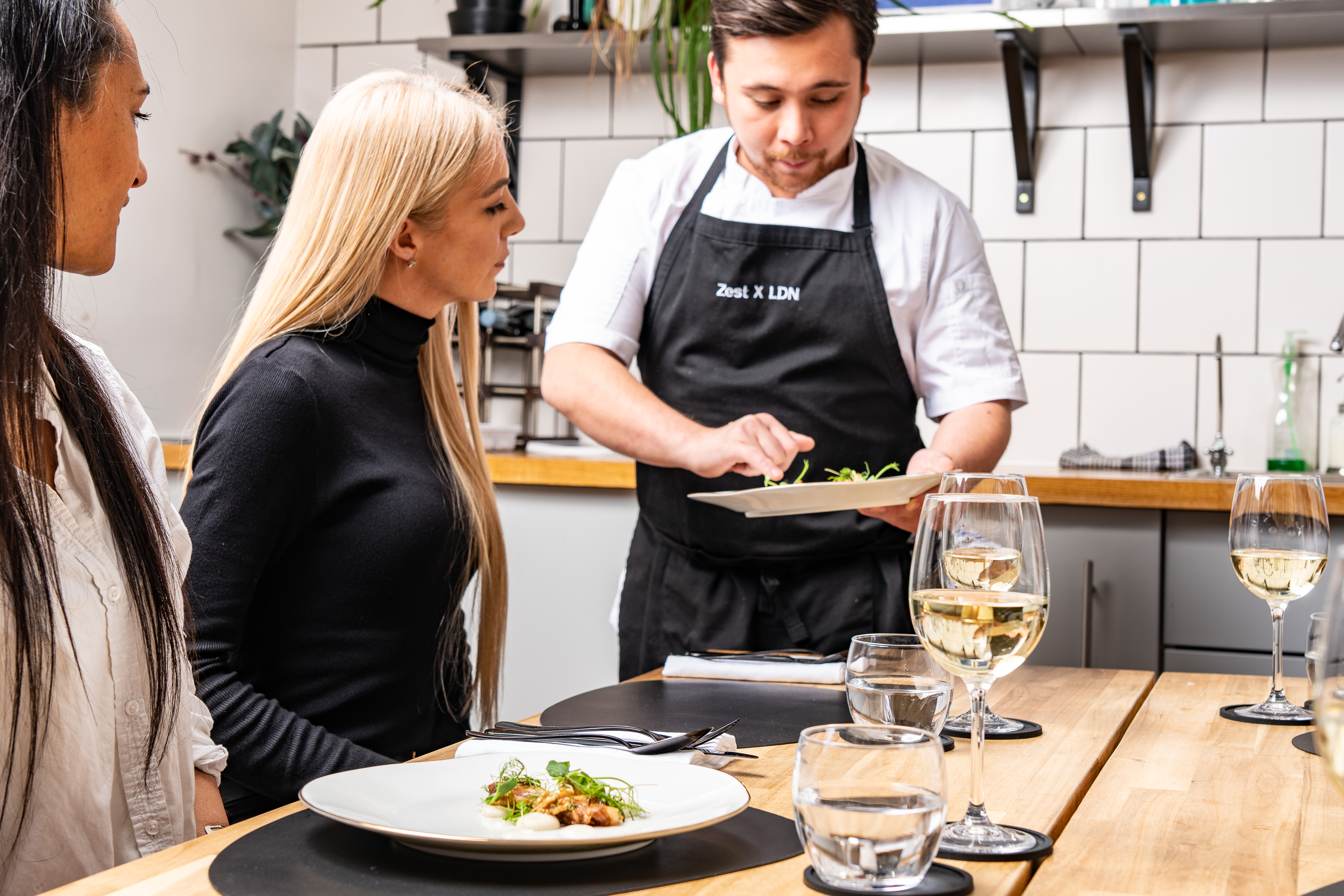 A chef in a black apron serves a beautifully plated dish to two attentive diners in a bright, minimalist space with wooden accents.