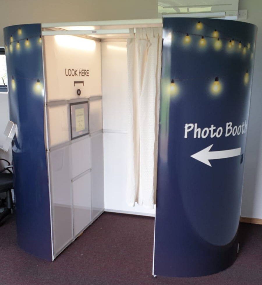 A modern photo booth with a navy exterior, illuminated by soft string lights, featuring a private curtain and easy-to-follow signage.