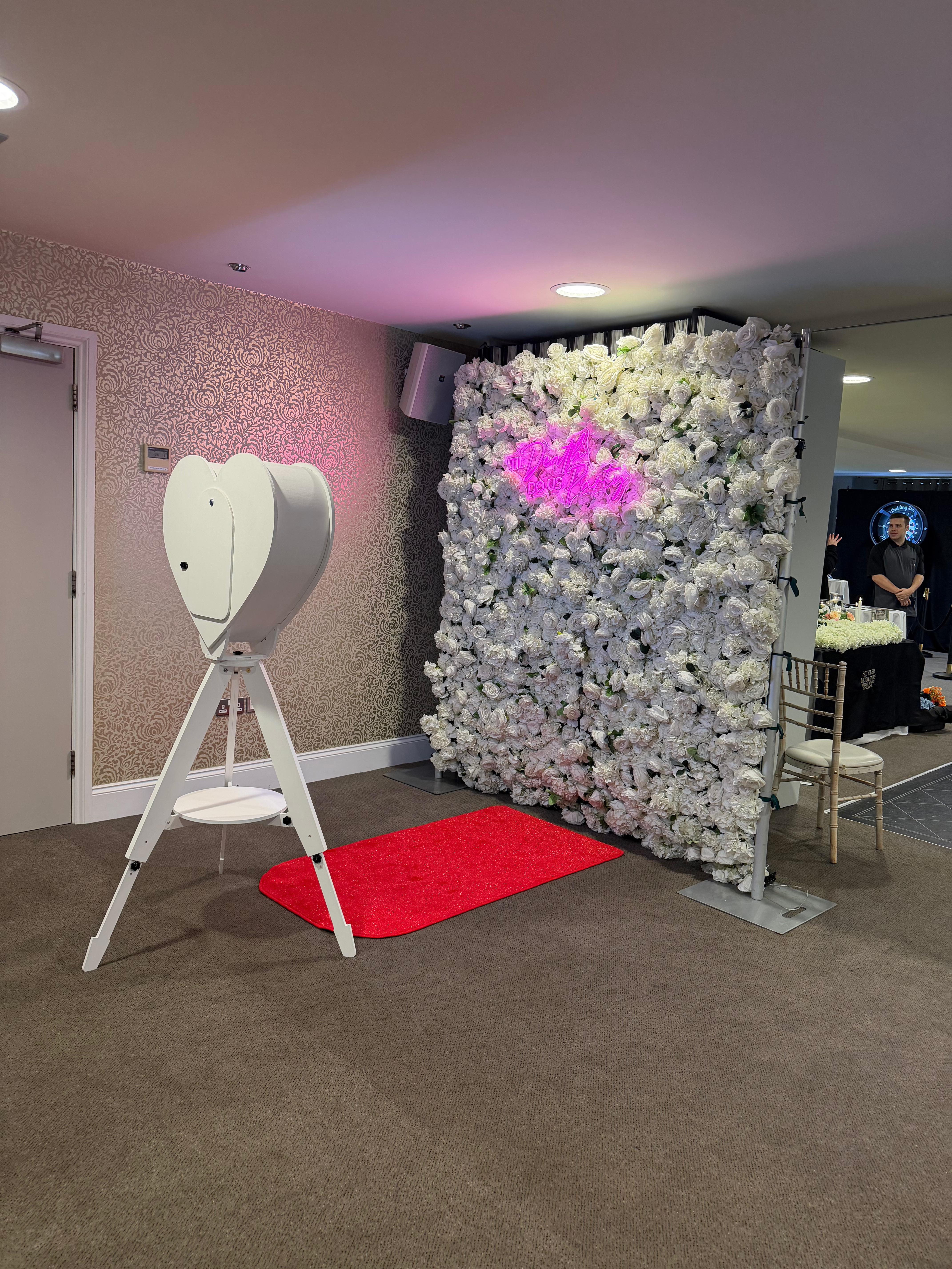 A white heart-shaped photo booth stands before a floral backdrop, adorned with lush white roses and a pink neon sign, creating an enchanting ambiance.
