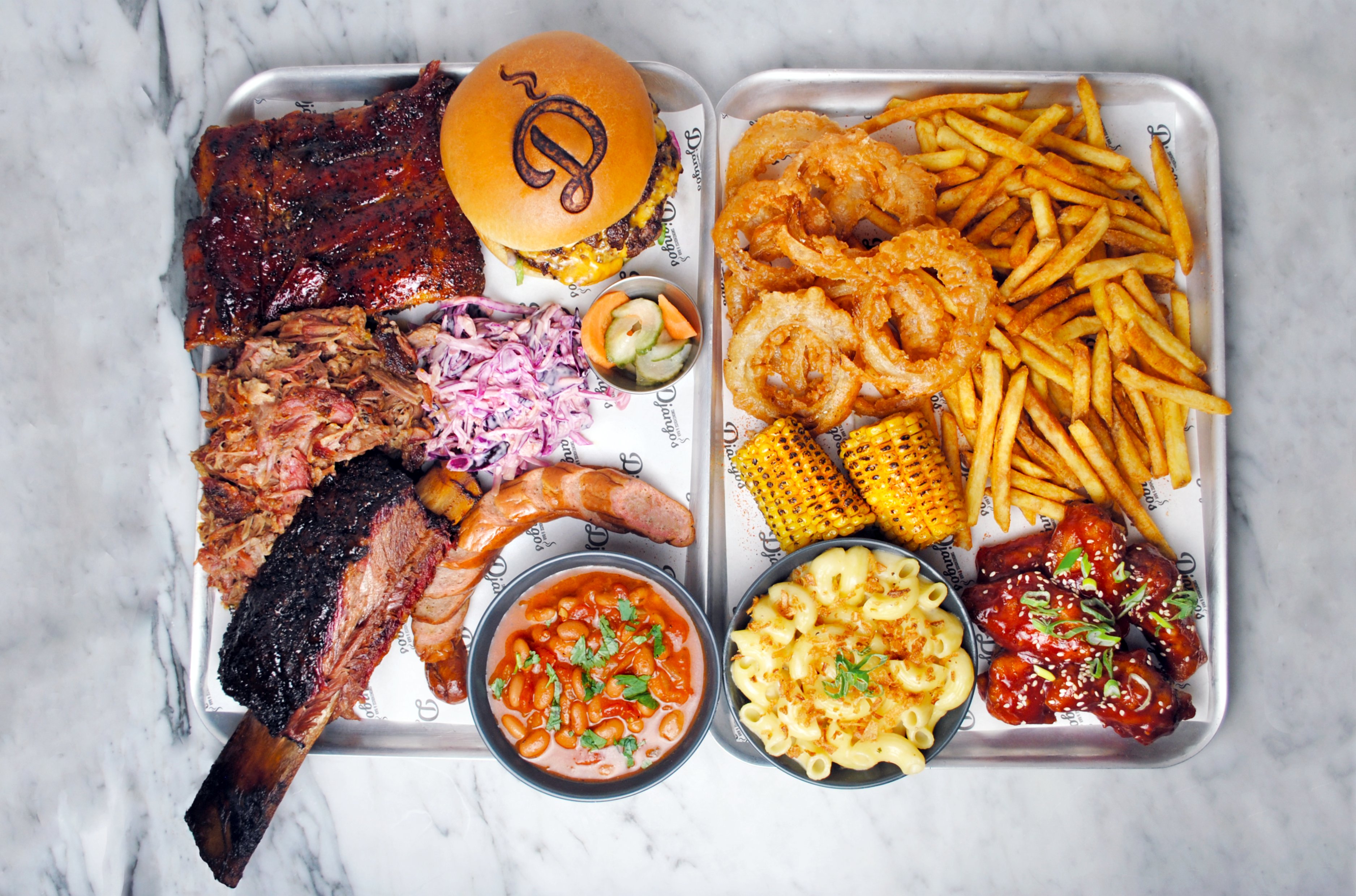Overhead BBQ catering tray on marble: ribs, brisket, pulled pork, burger, fries, onion rings, corn, mac and beans, glossy and artfully plated.