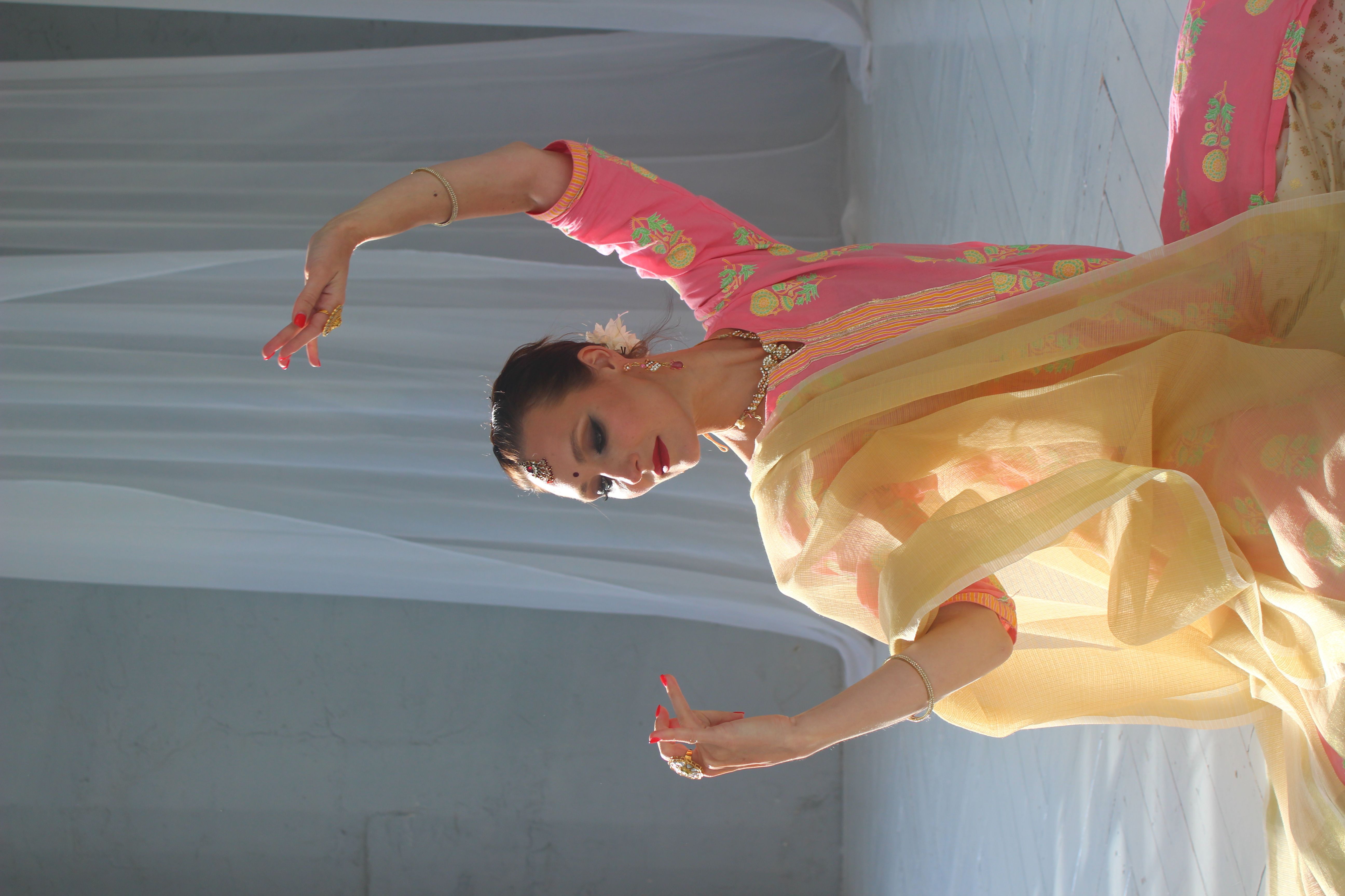 A dancer in a vibrant pink and yellow outfit gracefully poses against soft white drapes, illuminated by bright natural light, showcasing elegance.