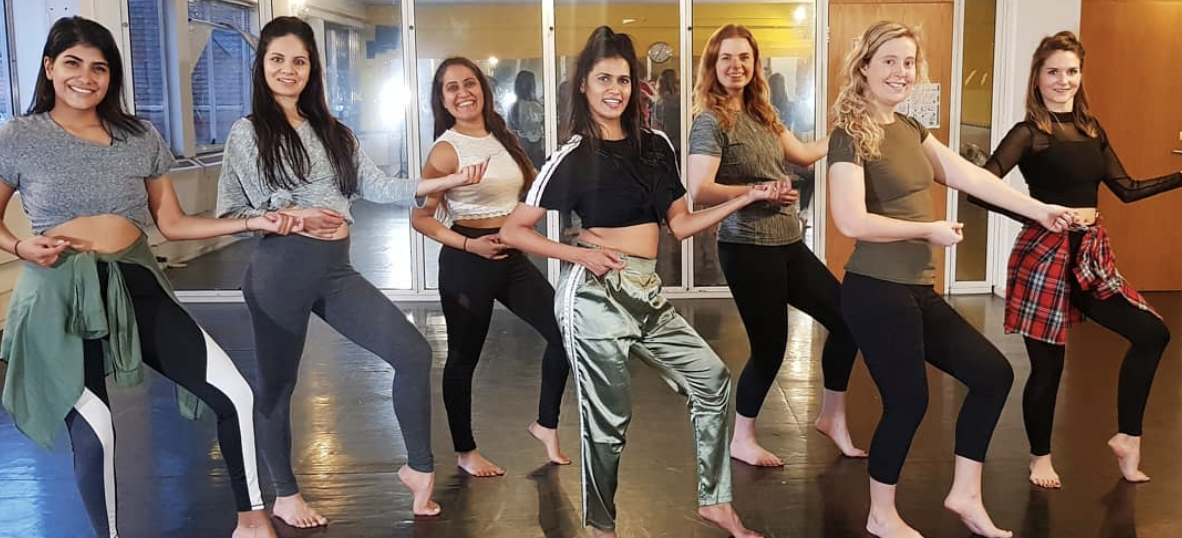 A vibrant dance studio filled with energy, featuring seven women in leggings and crop tops, joyfully striking poses in warm, natural light.