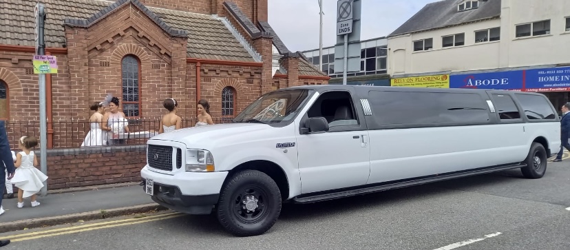 A sleek white limousine is parked outside a quaint brick church, while elegantly dressed bridesmaids in white gowns gather nearby, capturing moments of excitement.